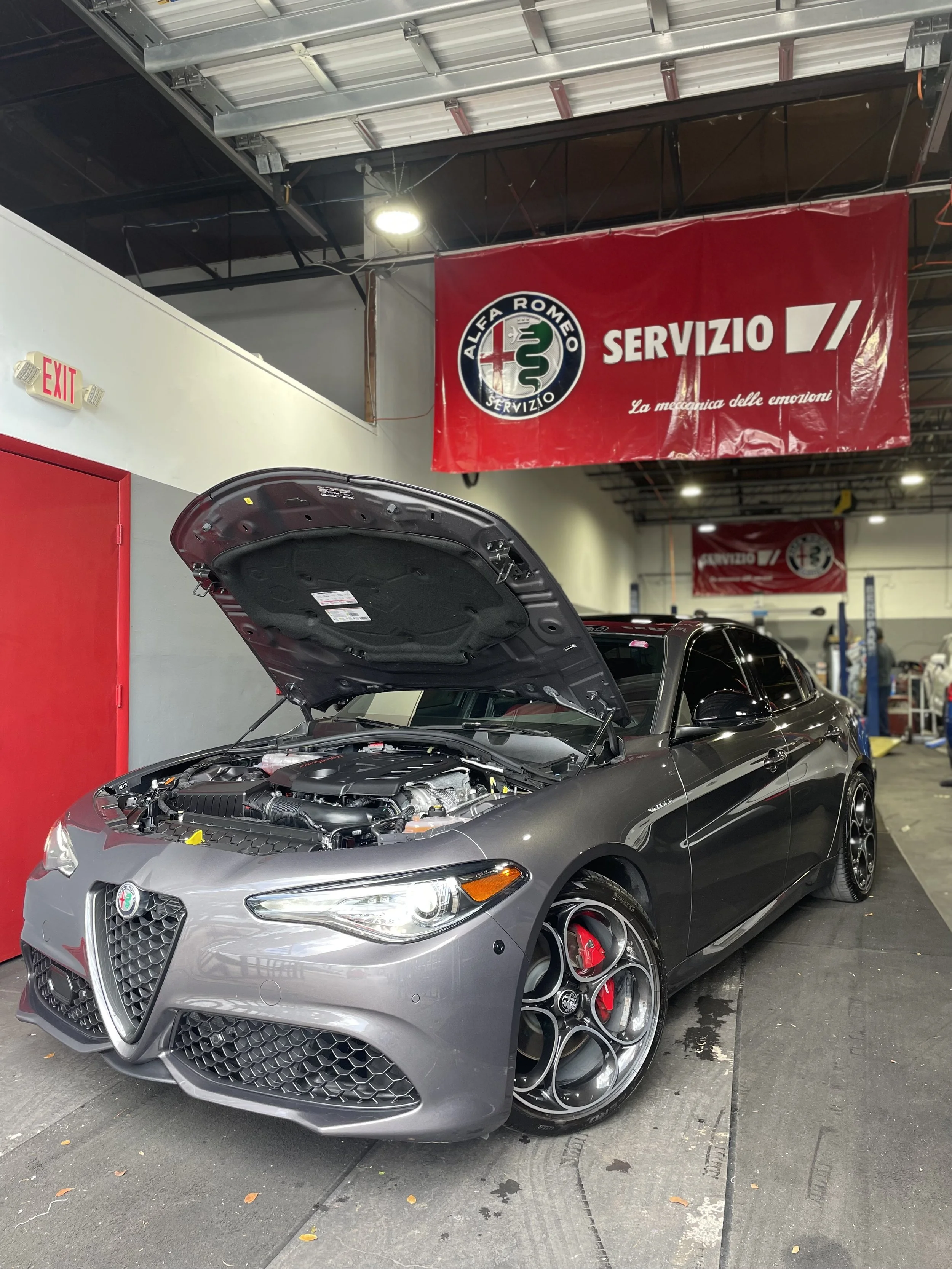 A silver Alfa Romeo car with its hood open, parked inside a service area. There is a red banner hanging from the ceiling that reads 'Alfa Romeo Servizio' and features a green serpent logo. The area has industrial ceiling lights and a red door on the side.