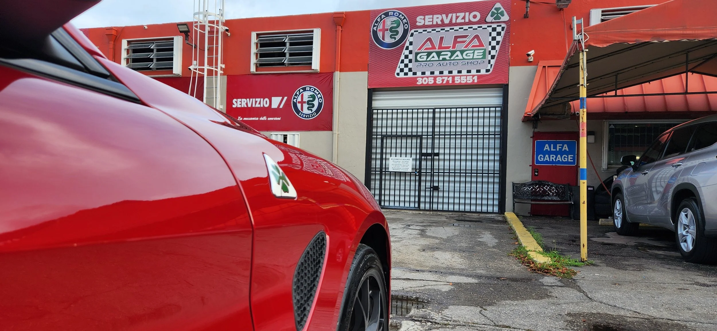 Red sports car parked outside Alfa Garage auto shop, with another car visible nearby. The garage features signs with Alfa Romeo branding and service advertisements, and there is a yellow parking barrier pole in front.