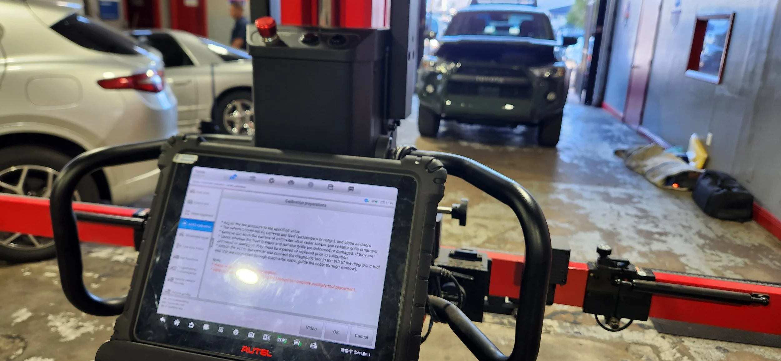 Inside an auto repair shop, with car diagnostic equipment and two vehicles on the lift, and a person working in the background.
