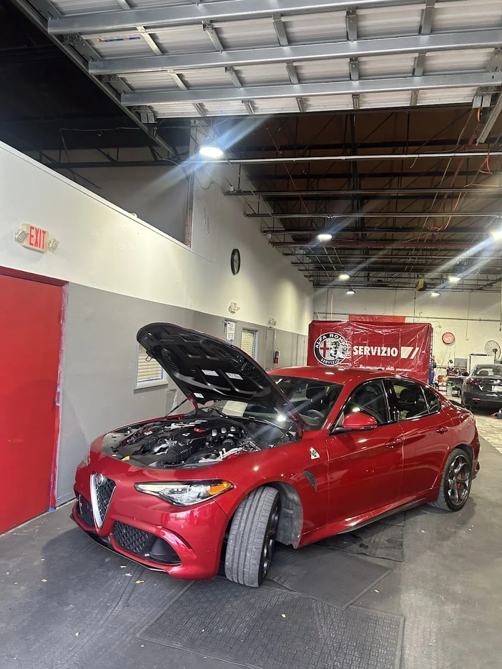Red Alfa Romeo sedan inside an auto repair shop with hood open and front left tire removed.
