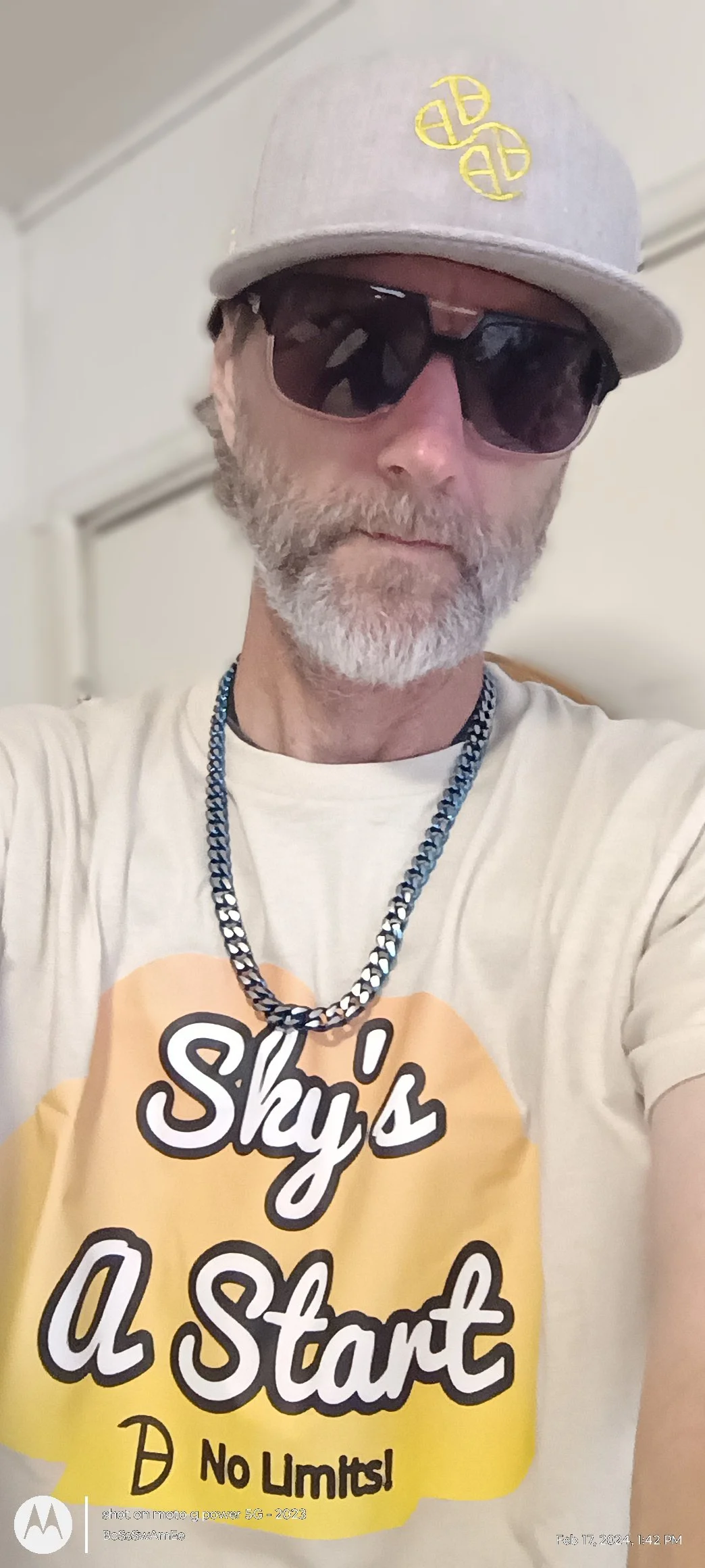 A man with a beard wearing a light gray cap with yellow seed logo, dark sunglasses, a white T-shirt with yellow text that says 'Sky's a Start' and 'No Limits,' a Blue colored Curb chain necklace, and standing.  bossswamee.com