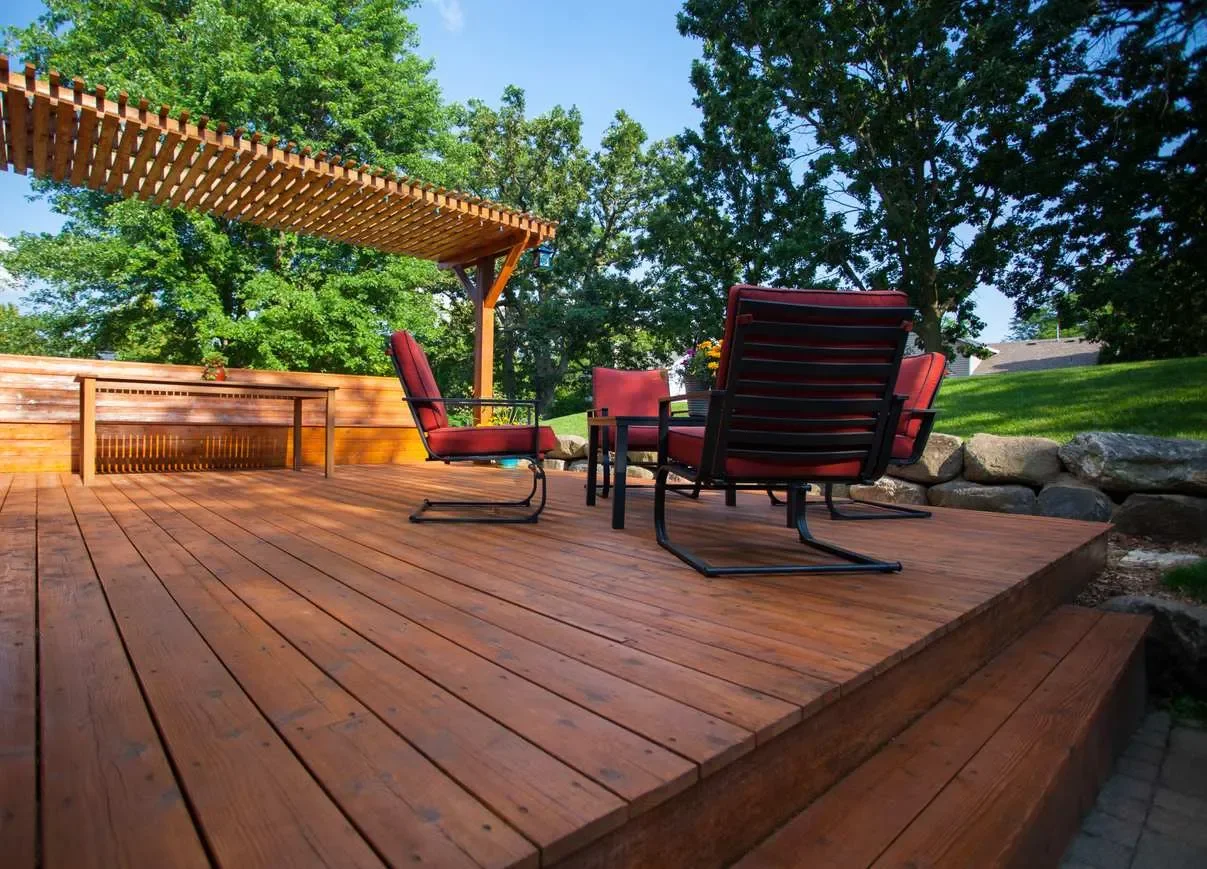 Terrasse en bois avec des chaises rouges et une pergola en bois, entourée d'arbres verts et d'un jardin.