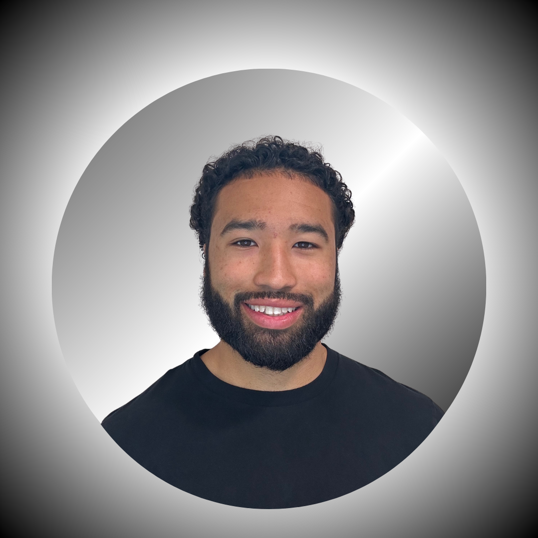 A young man with curly black hair and a beard smiling, wearing a black t-shirt, against a grey and white circular background.