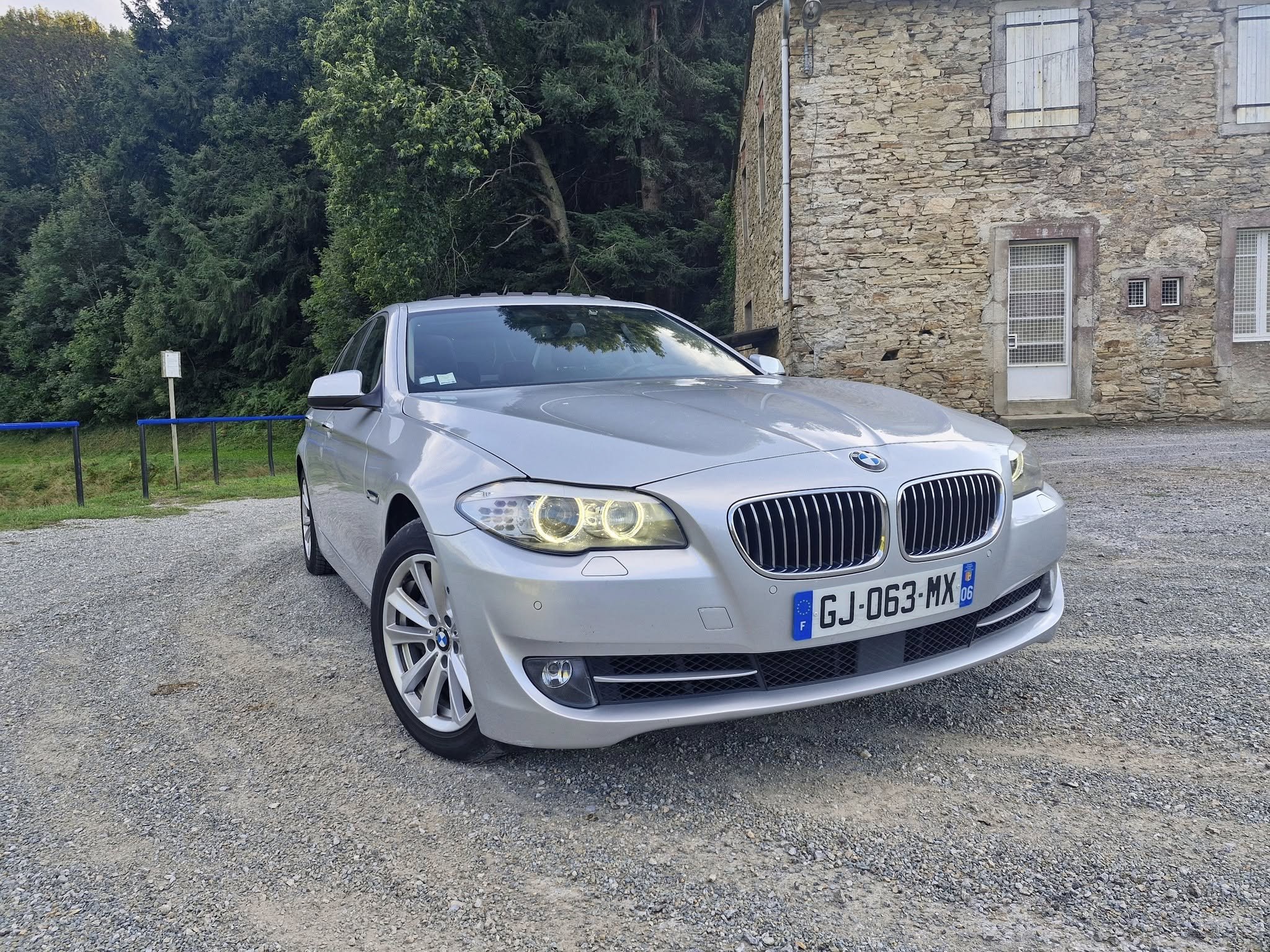 Voiture BMW grise stationnée sur un terrain en gravier devant un bâtiment en pierre avec des volets blancs.