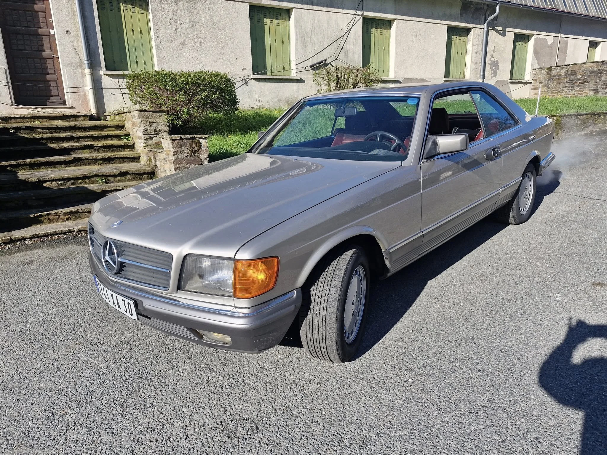 Voiture Mercedes-Benz ancienne argentée garée sur une rue, avec un bâtiment en arrière-plan et de l'herbe à côté.