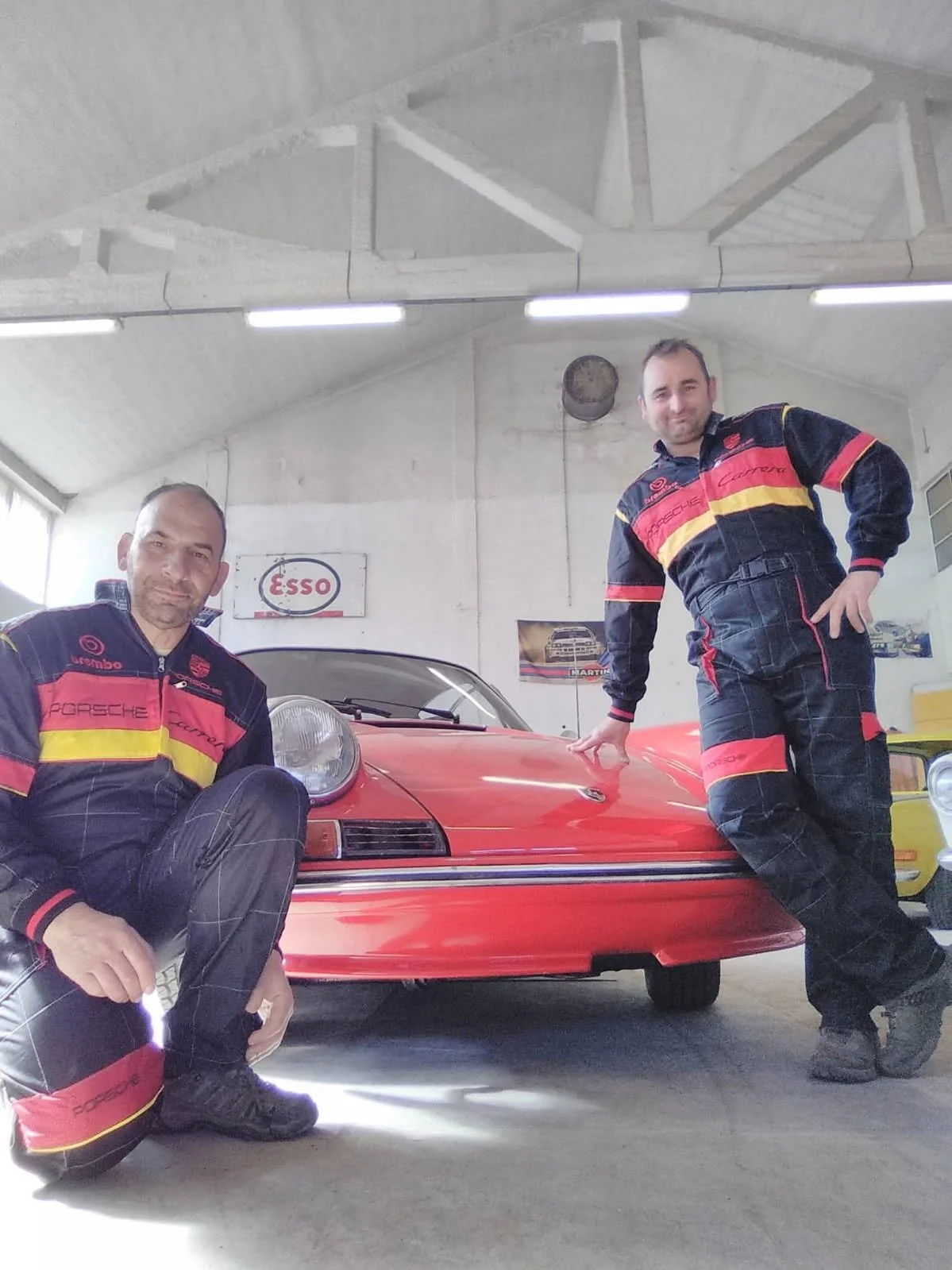 Deux hommes en combinaisons Porsche Carrera posent avec une voiture de collection Porsche, dans un atelier ou garage, avec des affiches et un panneau Esso au mur.
