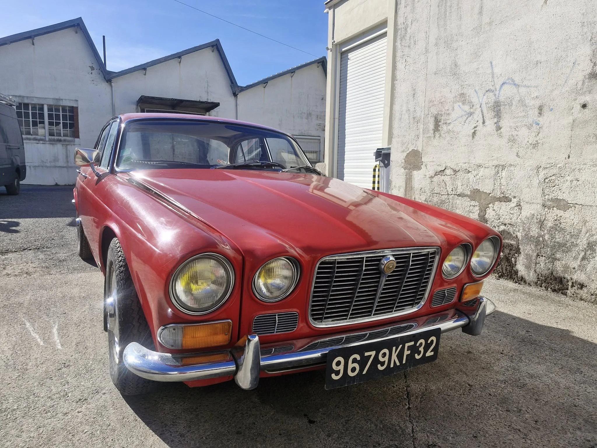 voiture rouge classique ancienne avec un design vintage, quatre phares ronds, calandre verticale et pare-chocs en chrome, stationnée devant un mur en béton