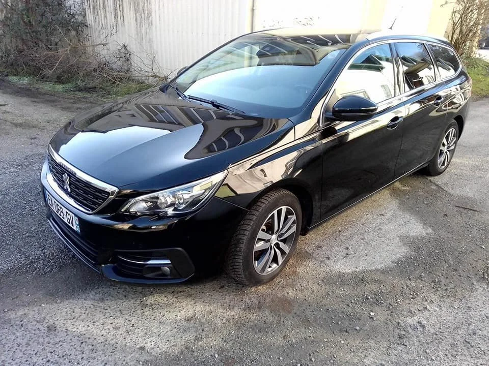 Voiture noire stationnée sur un chemin en extérieur, avec un bâtiment et des végétaux en arrière-plan.