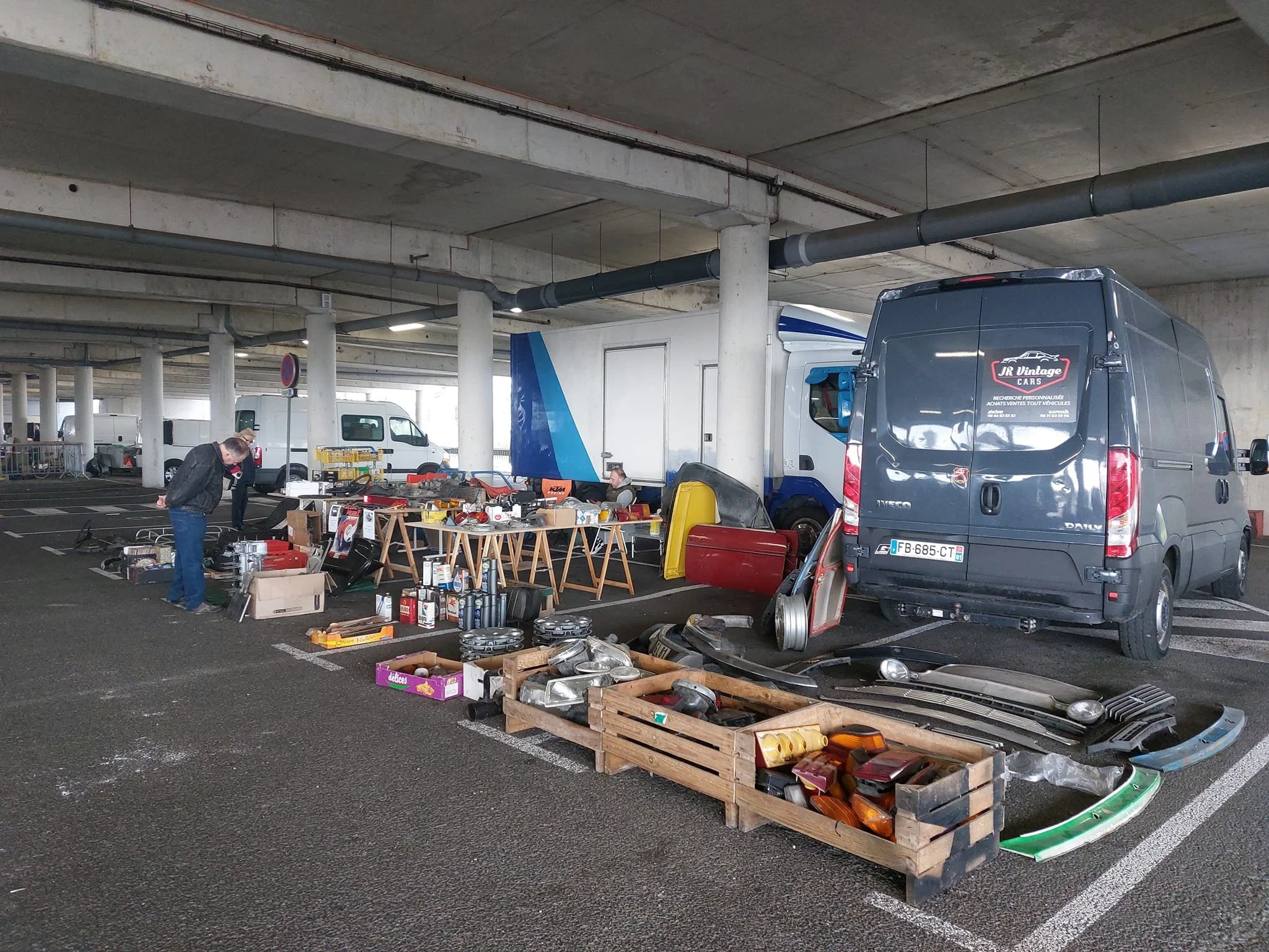 Vente de pièces détachées et objets d'occasion dans un parking, avec des voitures, des tables avec des objets divers, et des personnes examinant les articles.