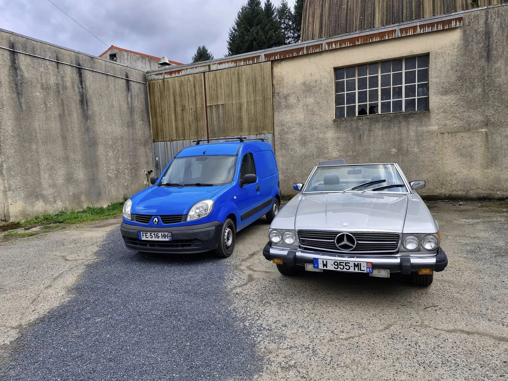 Une voiture bleue Renault Kangoo et une voiture décapotable Mercedes-Benz vintage argentée garées à côté d'un mur en béton dans une zone industrielle.