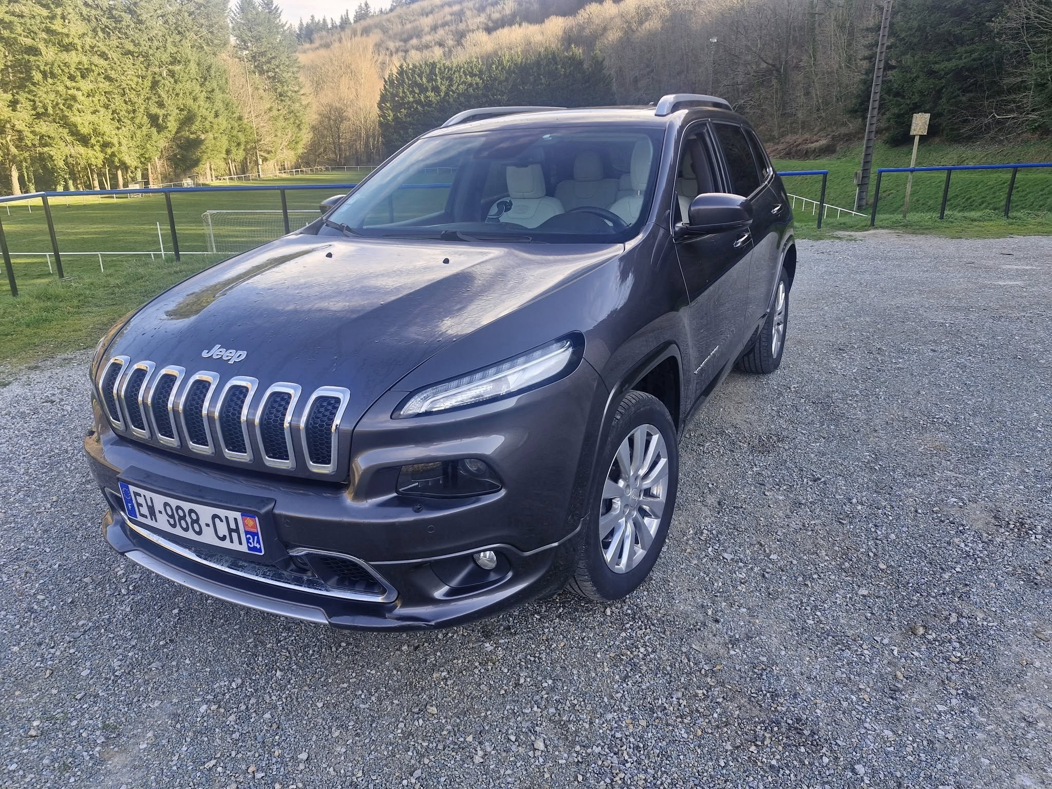 Voiture noire de modèle Jeep, stationnée sur un terrain en gravier, avec un environnement naturel comprenant des arbres et une pelouse en arrière-plan.