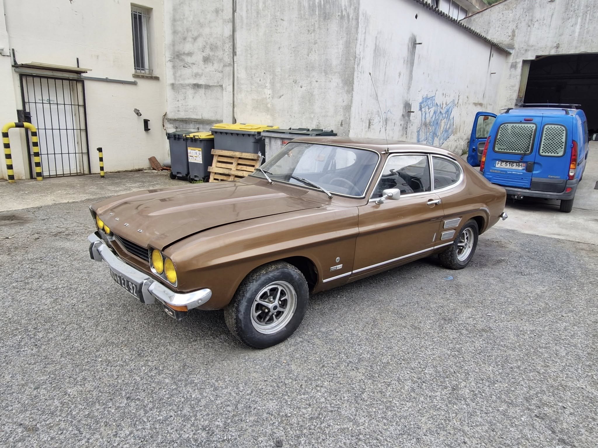 Voiture ancienne de couleur marron stationnée sur un parking en extérieur avec des poubelles et un bâtiment en fond.