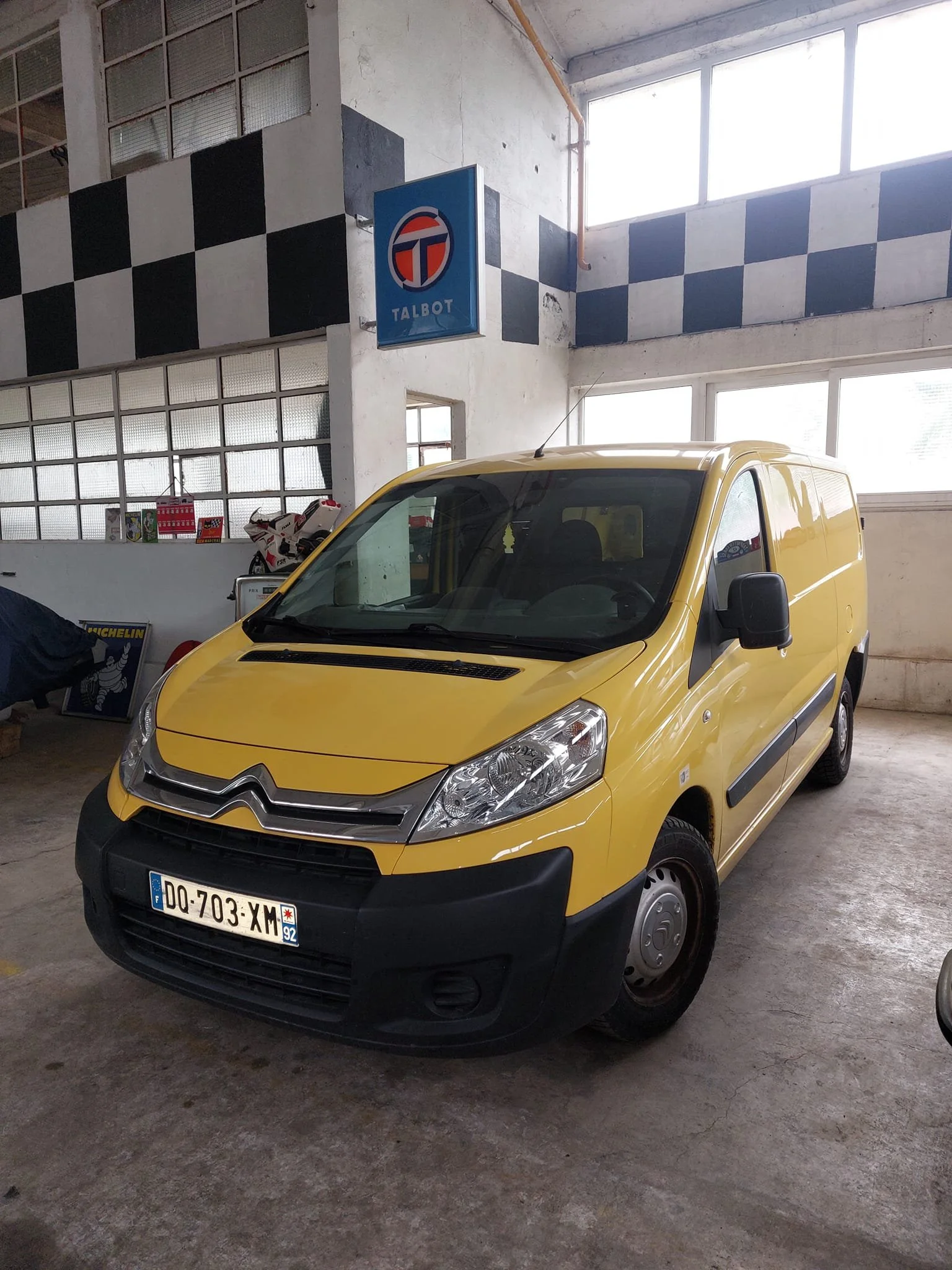 Une camionnette jaune stationnée dans un garage ou un atelier, avec un panneau Talbot en arrière-plan.