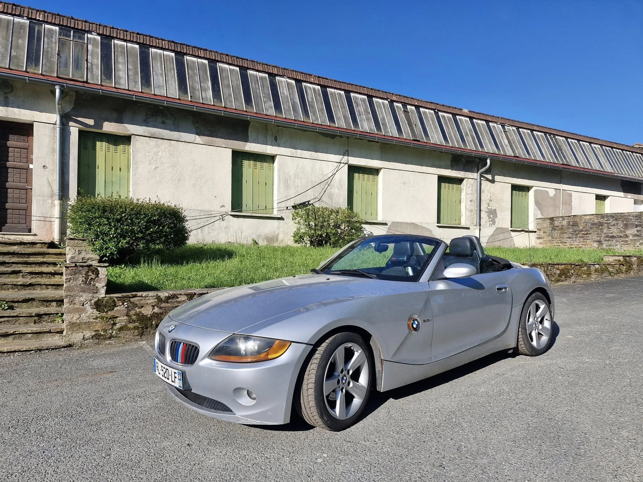 Voiture décapotable BMW grise garée devant une vieille maison avec volets verts, sous un ciel bleu.