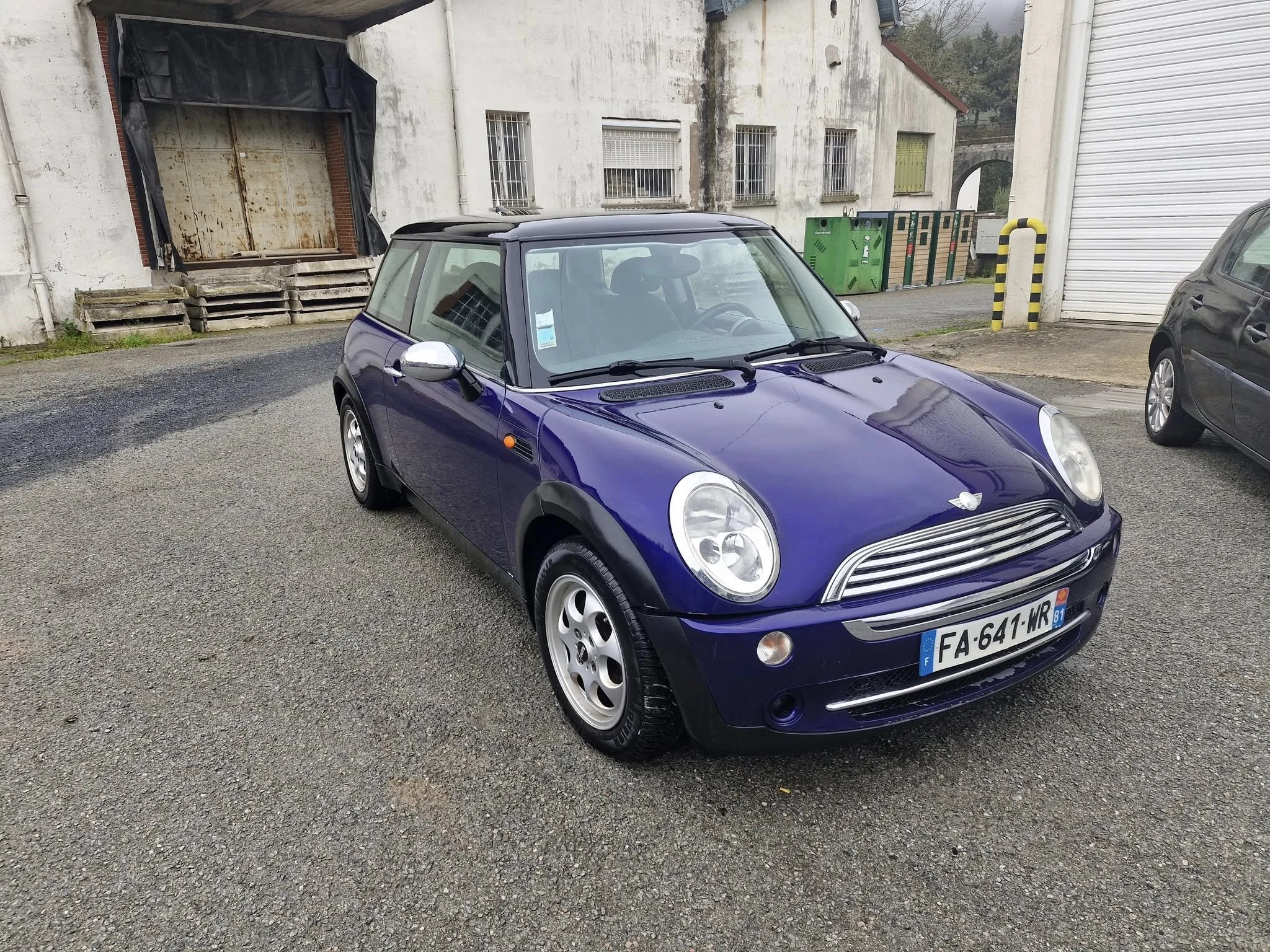 Voiture Mini Cooper violette stationnée dans un parking urbain avec des bâtiments anciens en arrière-plan.