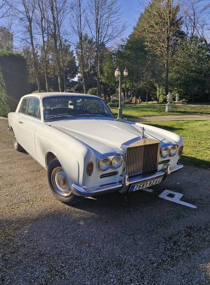 Voiture classique blanche, modèle de luxe, en stationnement à côté d'un parc avec des arbres et des lampadaires. Facilement reconnaissable par sa calandre et son design vintage.