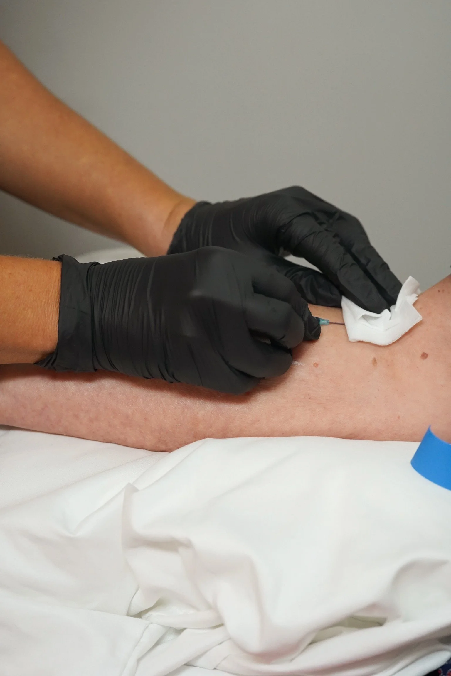 A healthcare professional wearing black gloves administers an injection into a patient's leg using a syringe and a cotton ball for pressure.