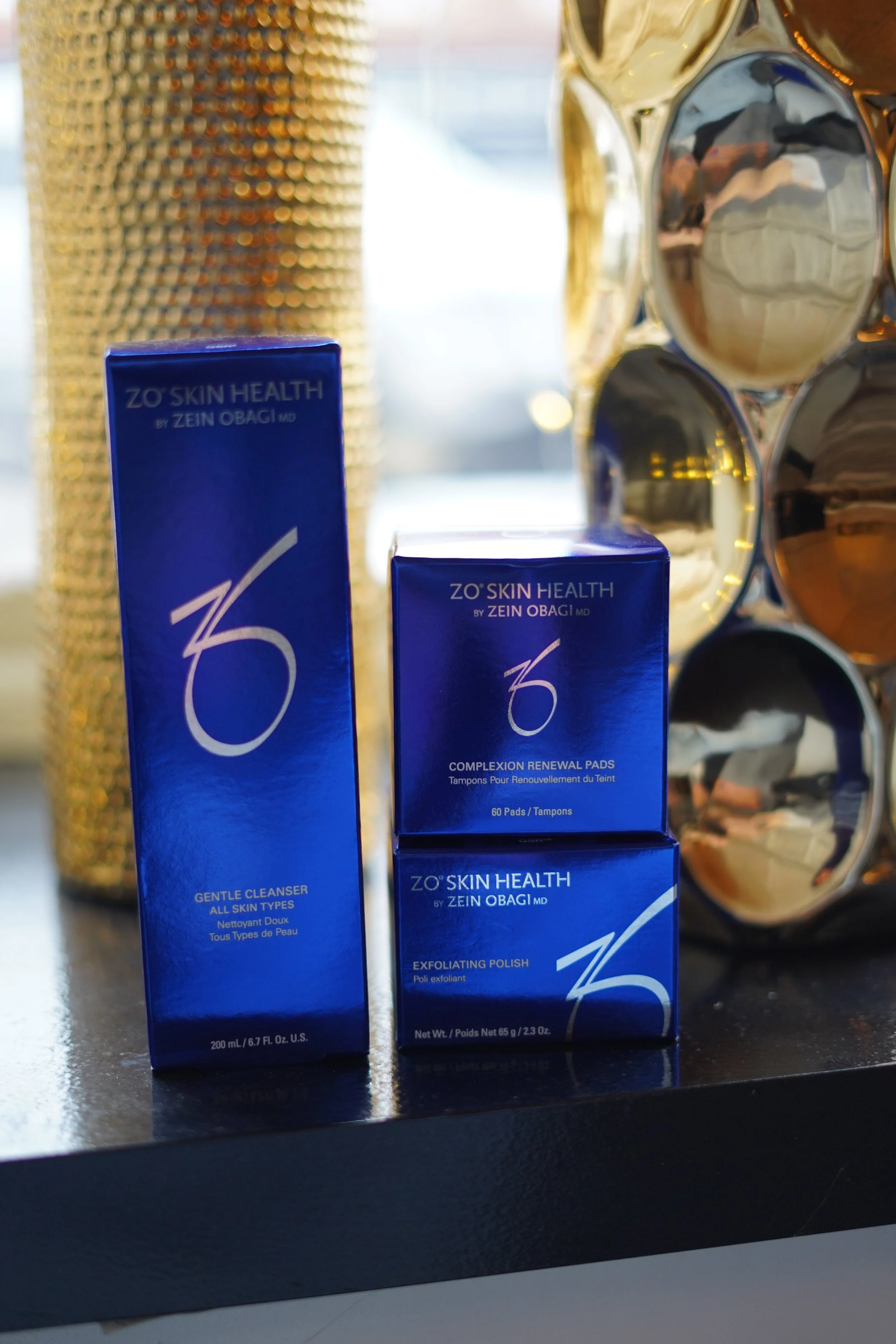 Three skincare products from ZO Skin Health, including gentle cleanser, exfoliating polish, and renewal pads, placed on a black surface with decorative gold and silver objects in the background.