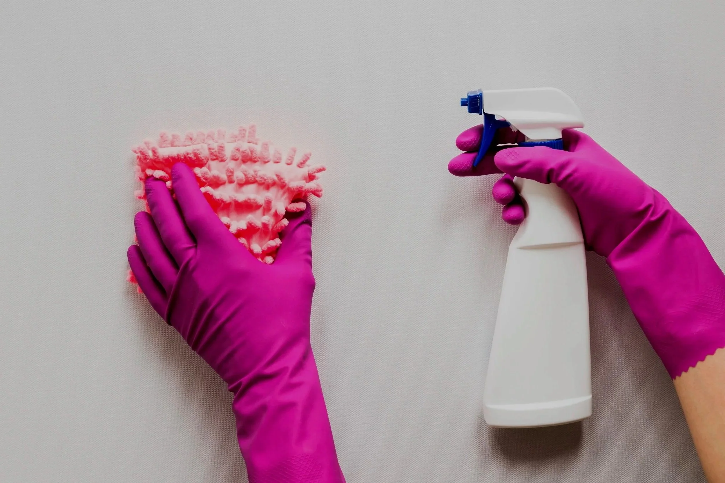 Hands wearing pink rubber gloves cleaning a white surface with a pink cleaning cloth and a white spray bottle.