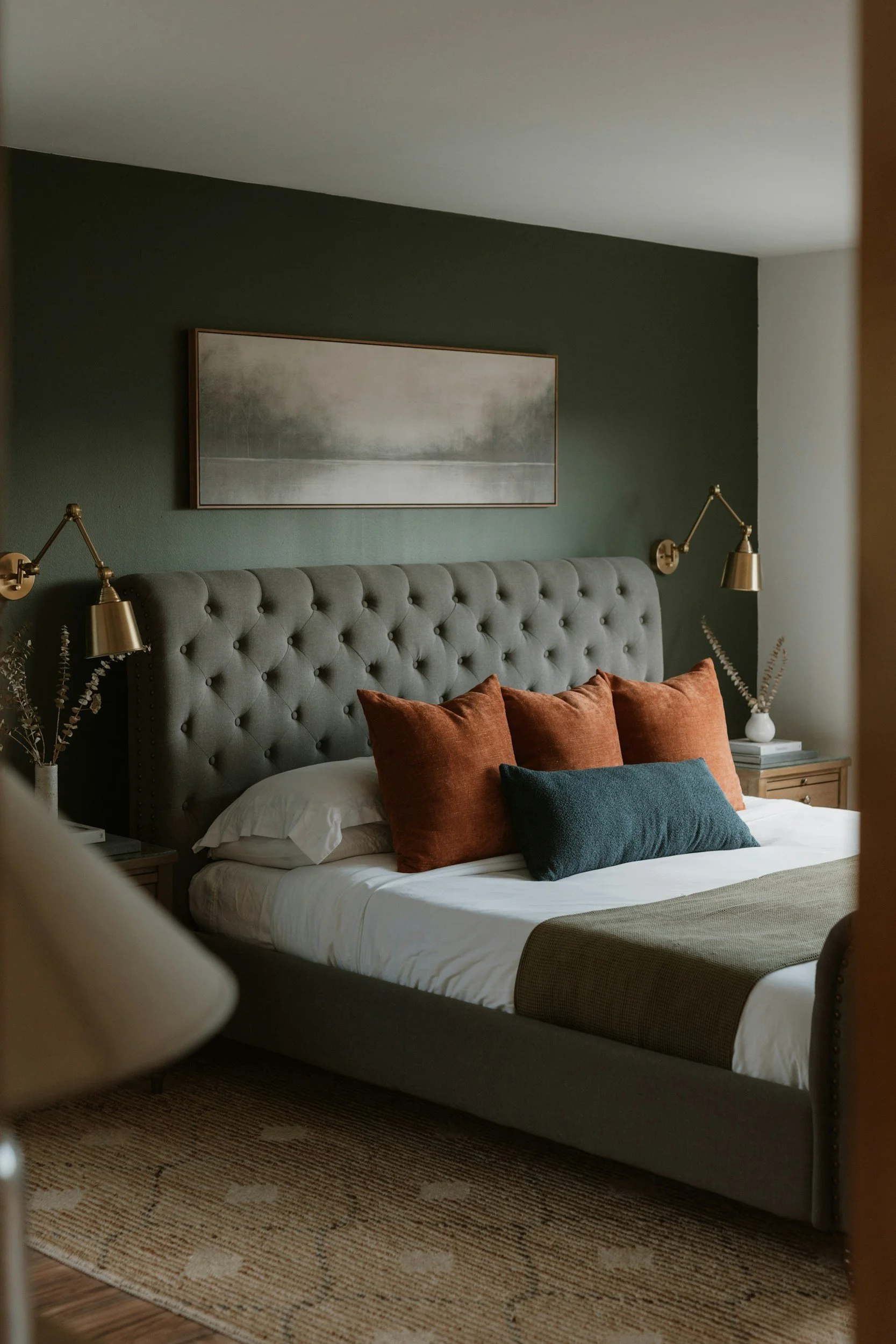 A cozy bedroom with a tufted gray headboard, green accent wall, and gold wall-mounted reading lamps. The bed has white sheets, four rust-colored pillows, and one dark blue pillow. There are two nightstands with white vases and books, and a framed landscape painting above the bed.