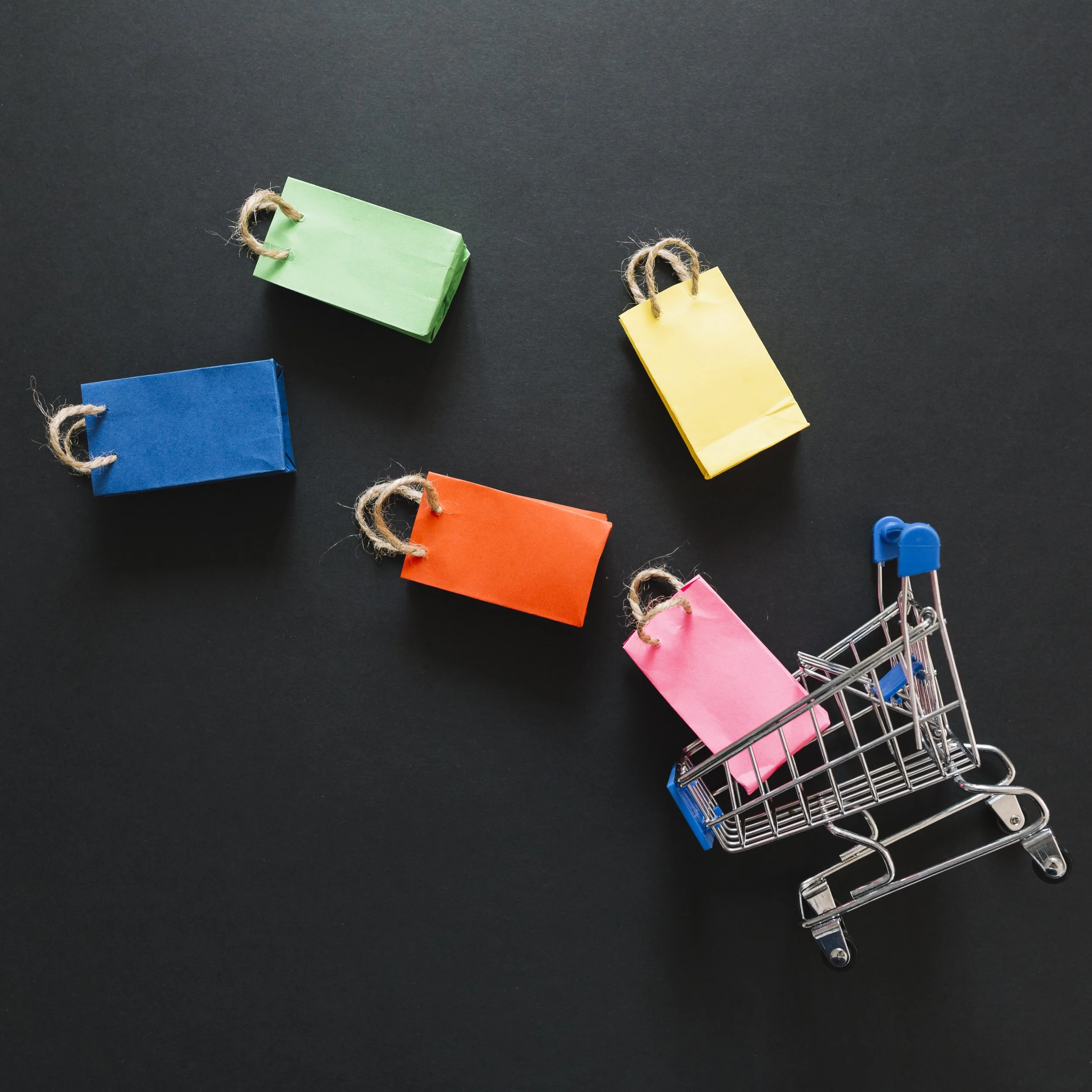Miniature shopping cart with pink paper tag inside, surrounded by colored paper tags in green, blue, orange, and yellow on a black background.