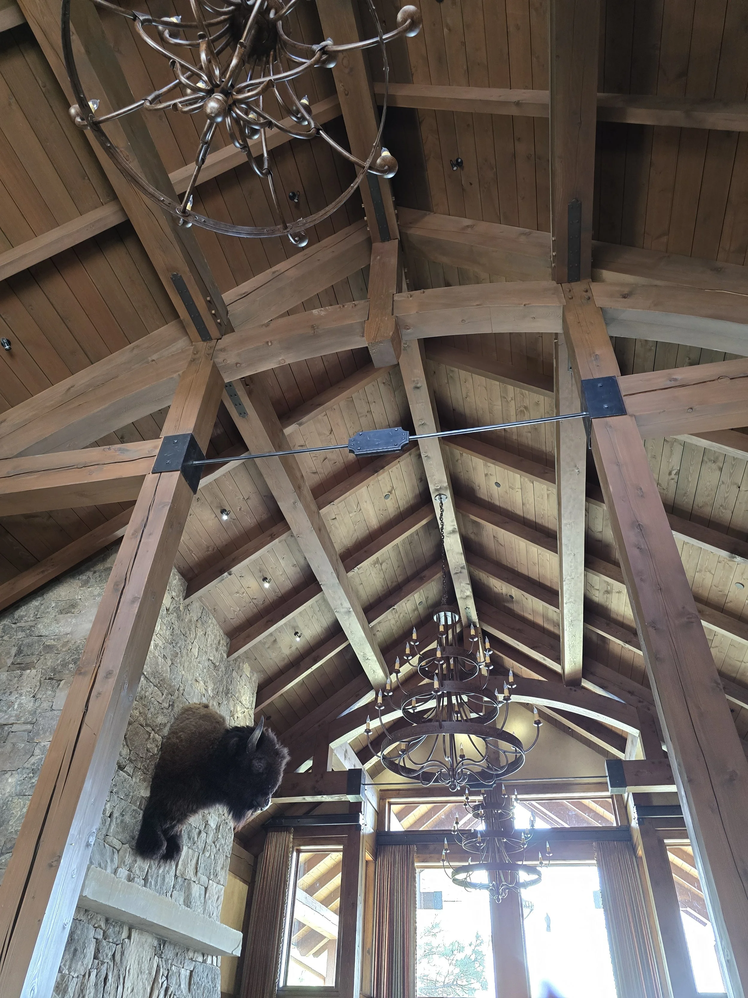 Interior view of a cathedral-like room with wooden beams, chandeliers, a stone fireplace with a mounted animal head, and large windows with curtains.