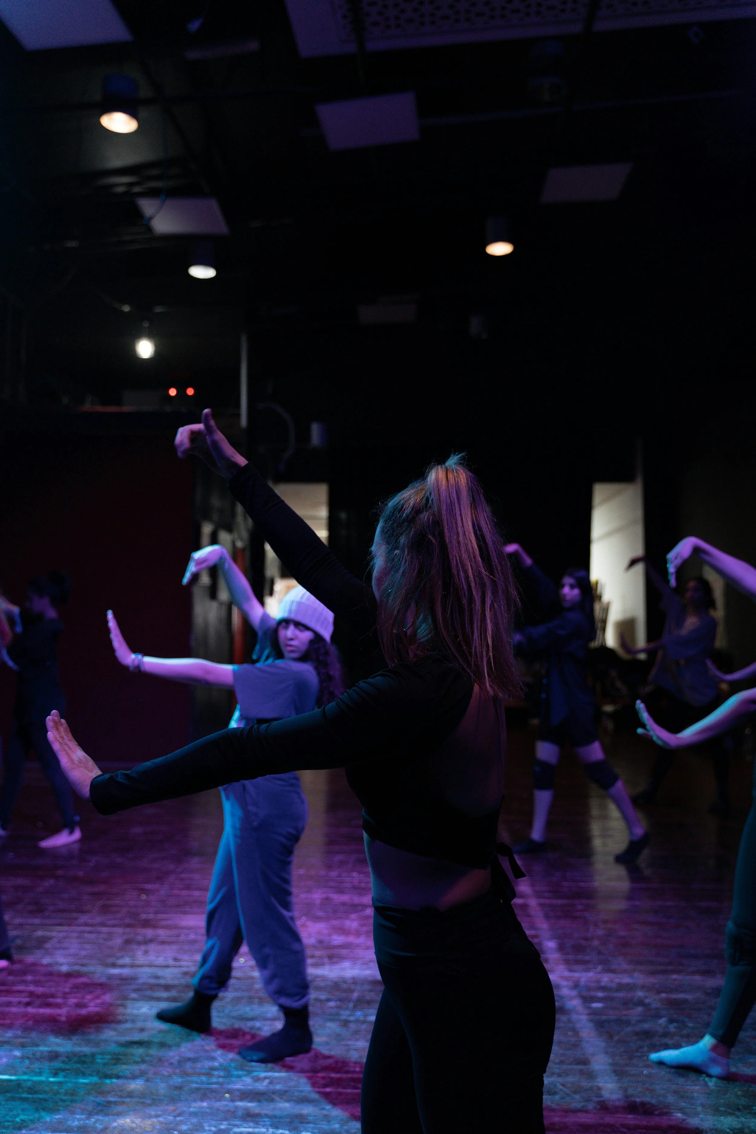 A group of dancers rehearsing in a dimly lit studio with colorful lighting, some wearing casual and comfortable clothing.