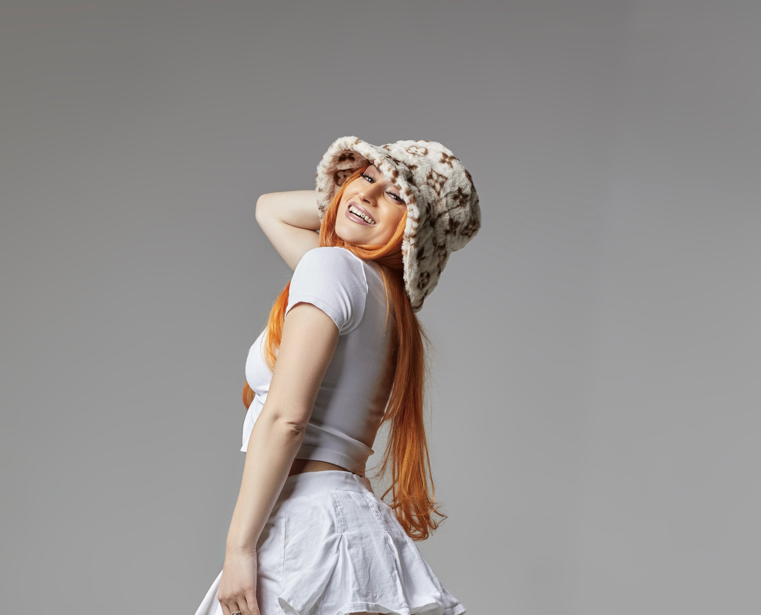 Woman with long red hair smiling, wearing a fuzzy hat and a white outfit, standing against a plain gray background.