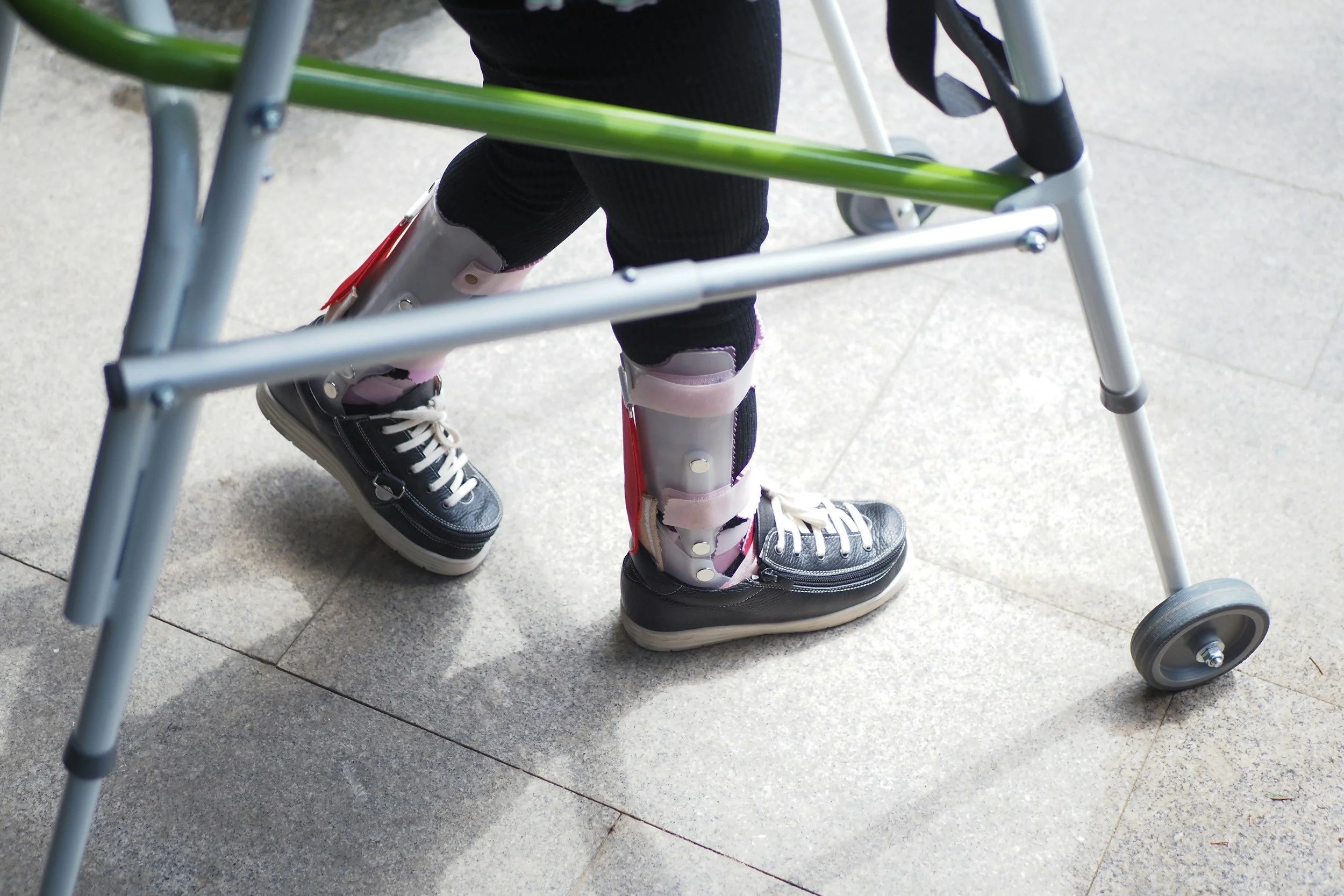 A person wearing black sneakers with white laces and black pants using a walker with medical ankle braces on both ankles.