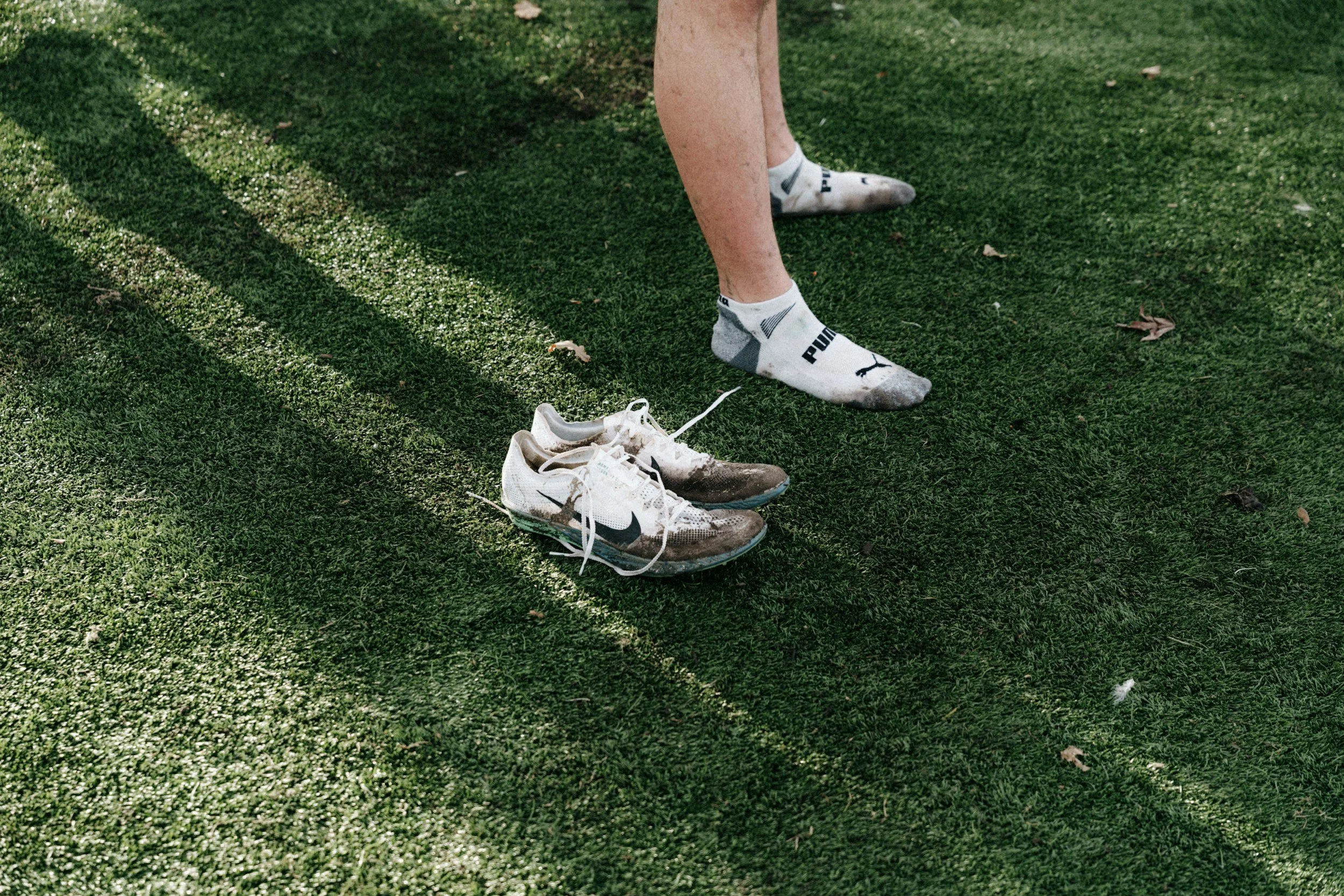 A person standing on grass near a pair of muddy running shoes with one white sock visible.