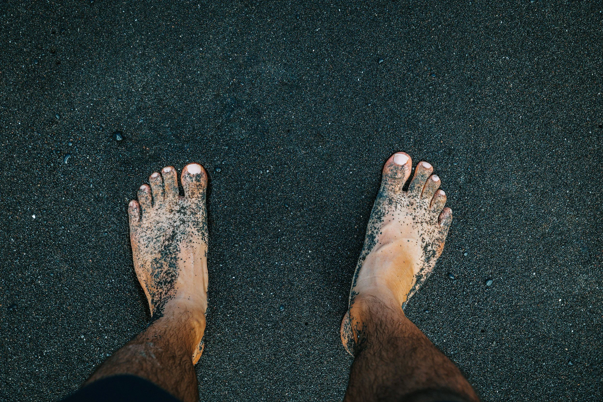 Feet standing in wet, black sand on a beach with sand sticking to the toes and legs.