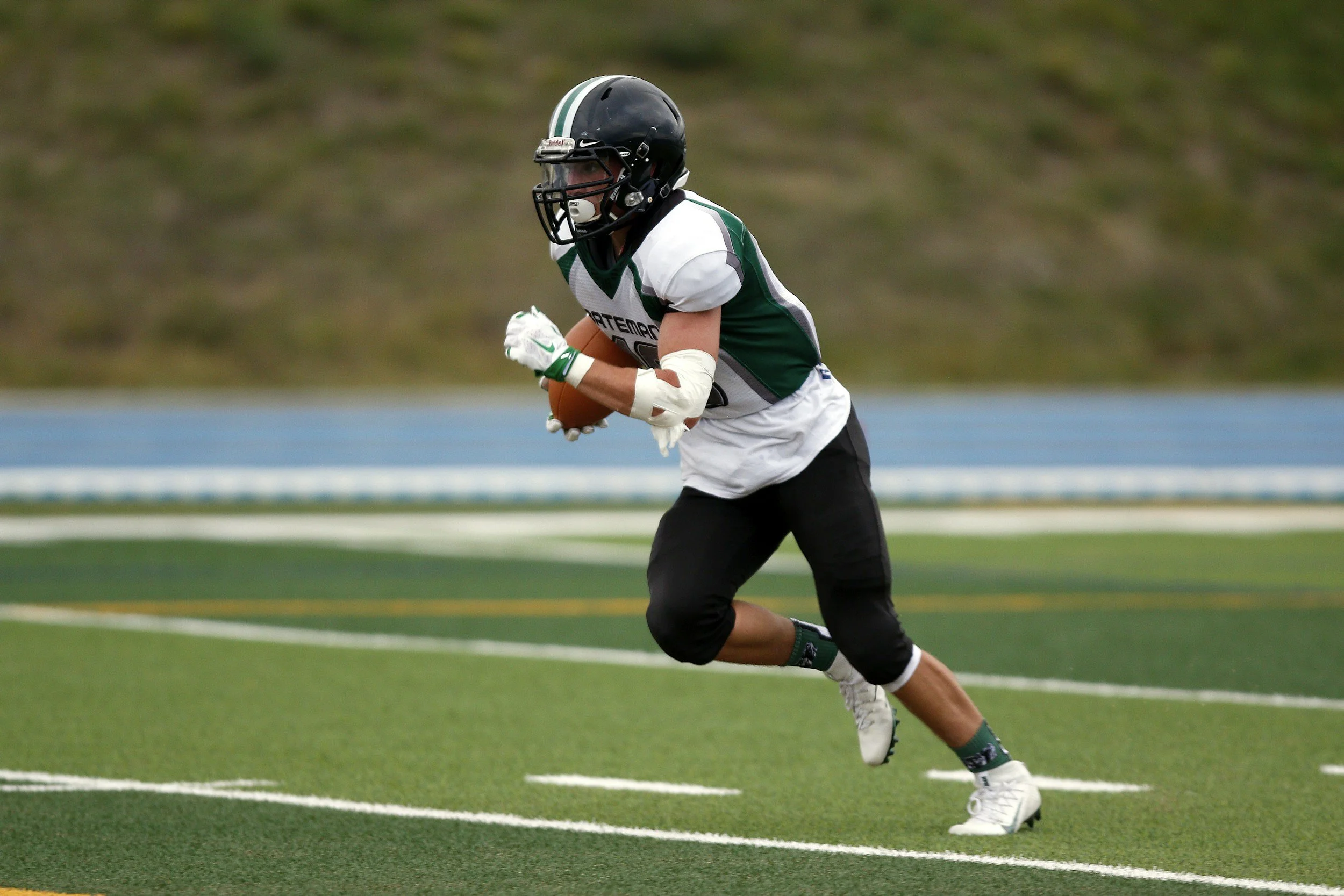 Football player in a green and white uniform holding a football while running on the field.
