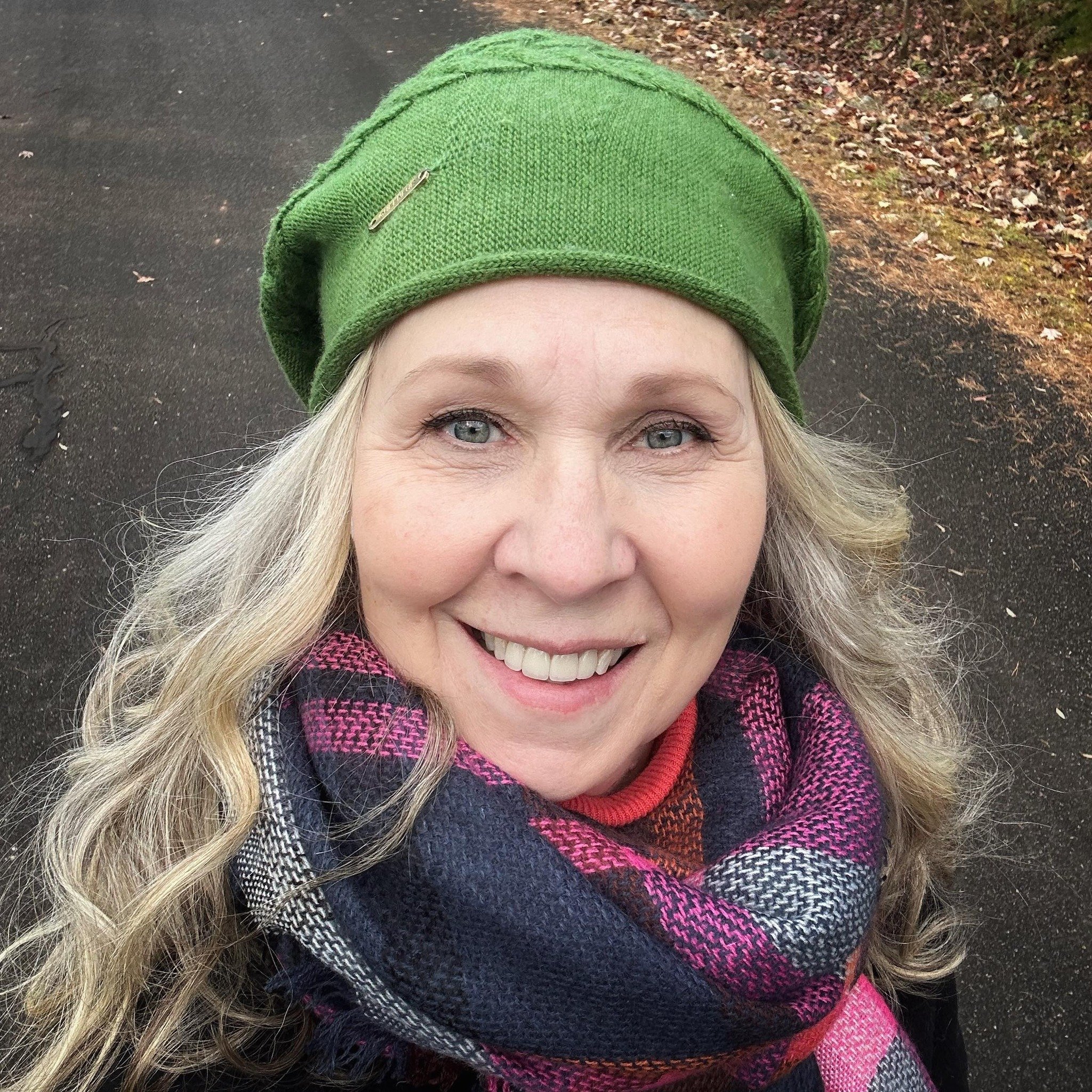 Dolores Chandler has long blonde hair, blue eyes, and a big smile, wearing a green knit hat and a multicolored plaid scarf, standing on a paved path with fallen leaves on the ground.