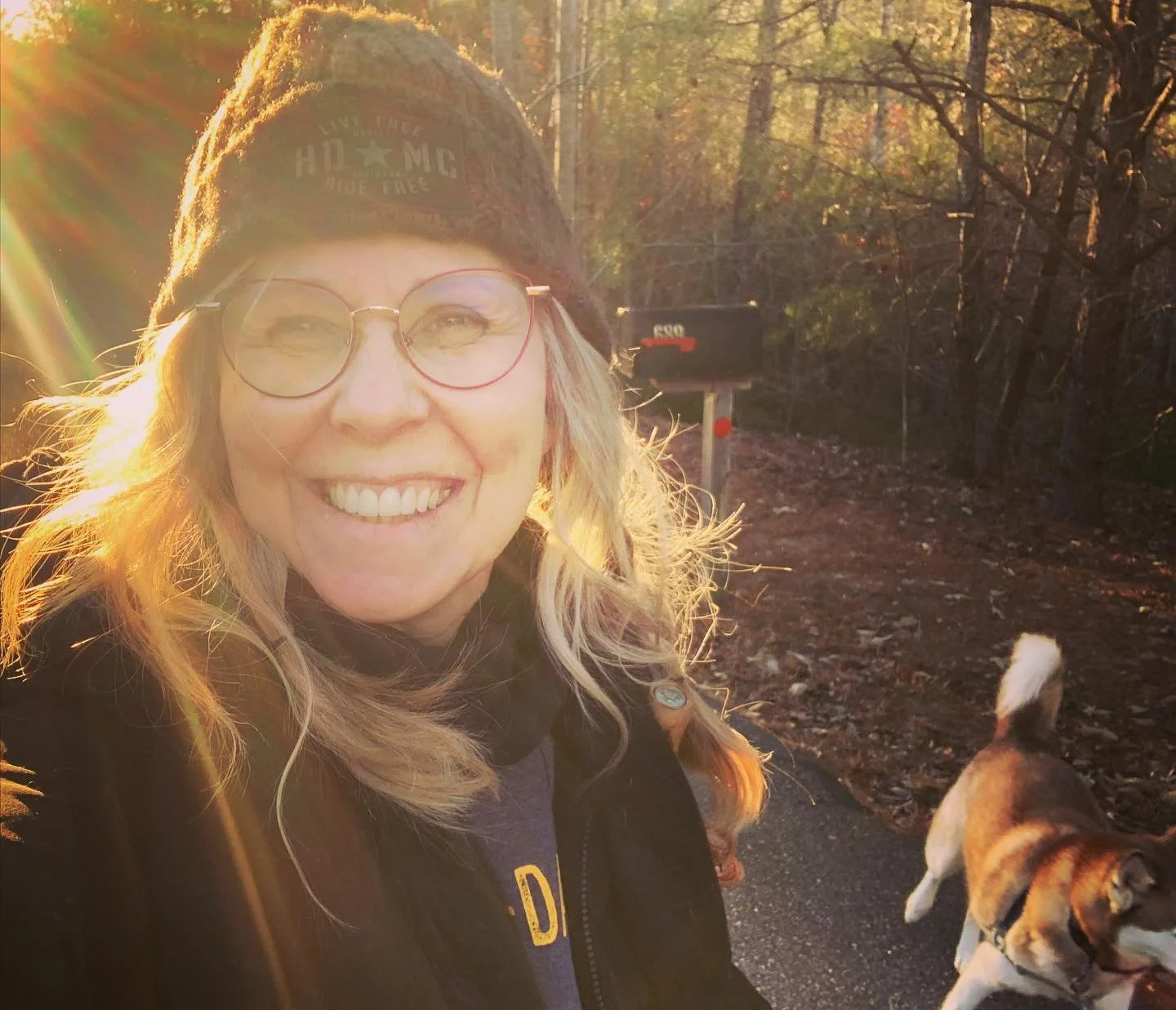 Dolores Chandler is smiling outdoors during sunset, wearing glasses, a knit beanie, and a dark jacket, with a dog walking behind her on a wooded trail.