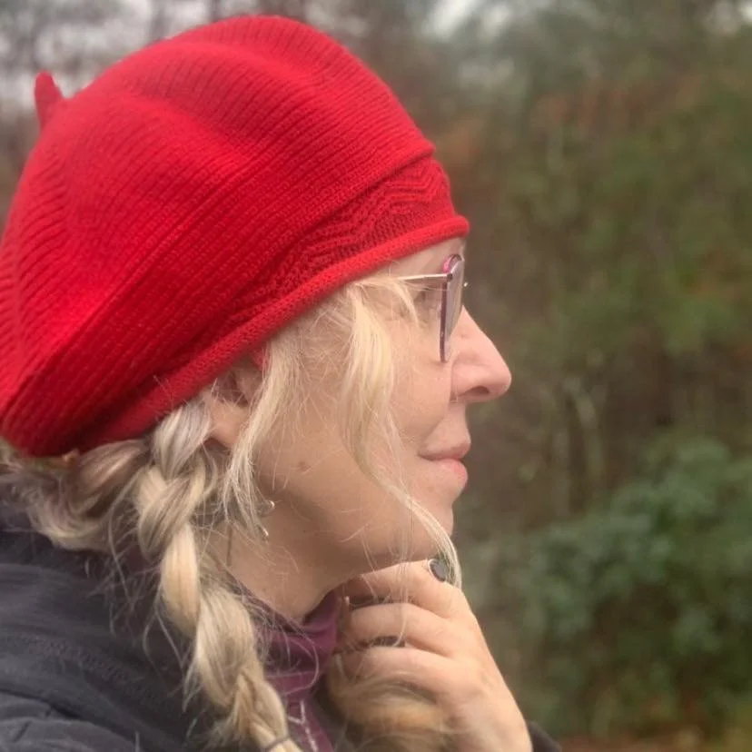 Dolores Chandler has blonde hair in a braid wearing glasses and a red knit hat outdoors.