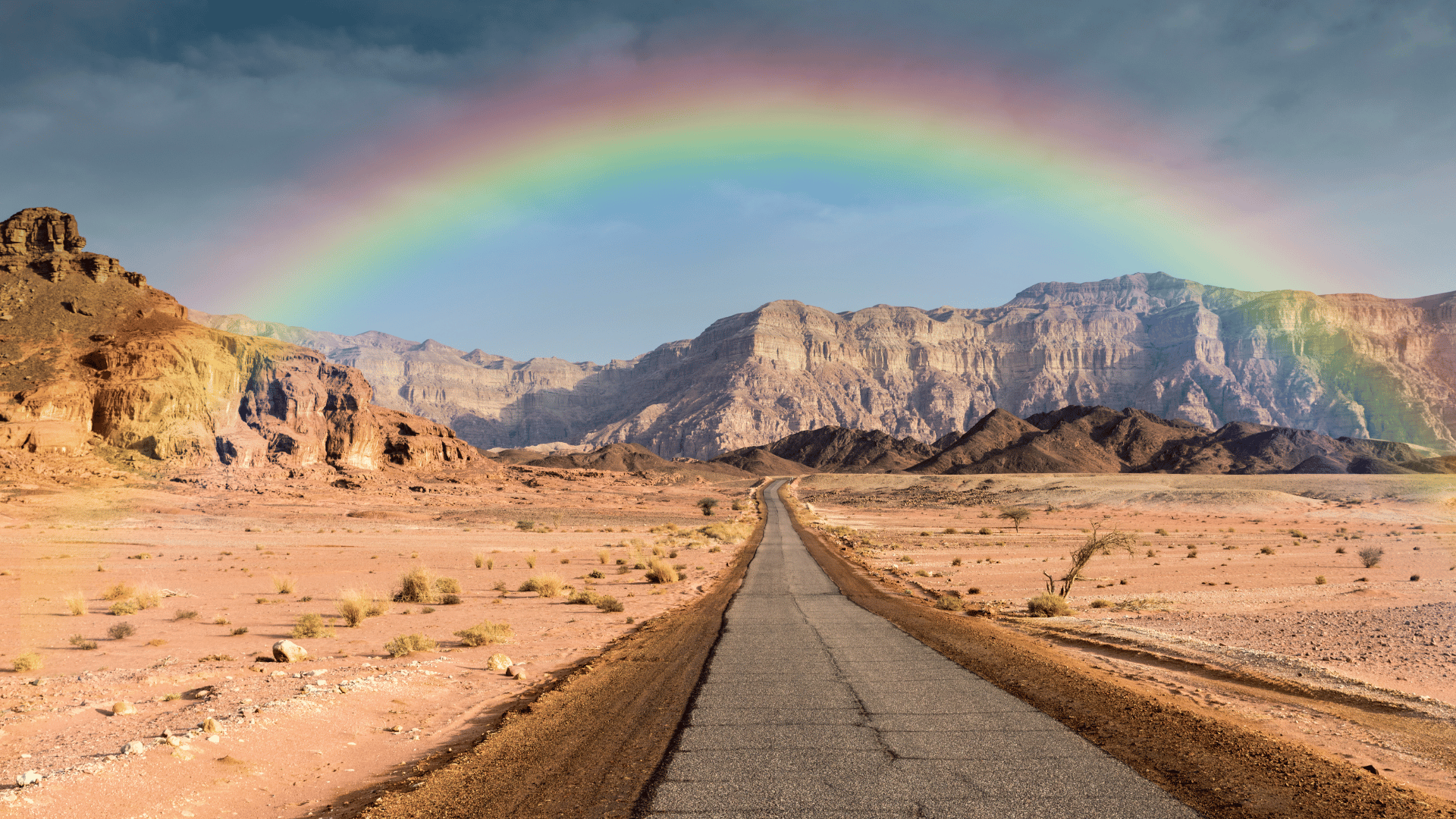 A long, straight road running through a desert landscape with rocky mountains and a rainbow arching across the sky.