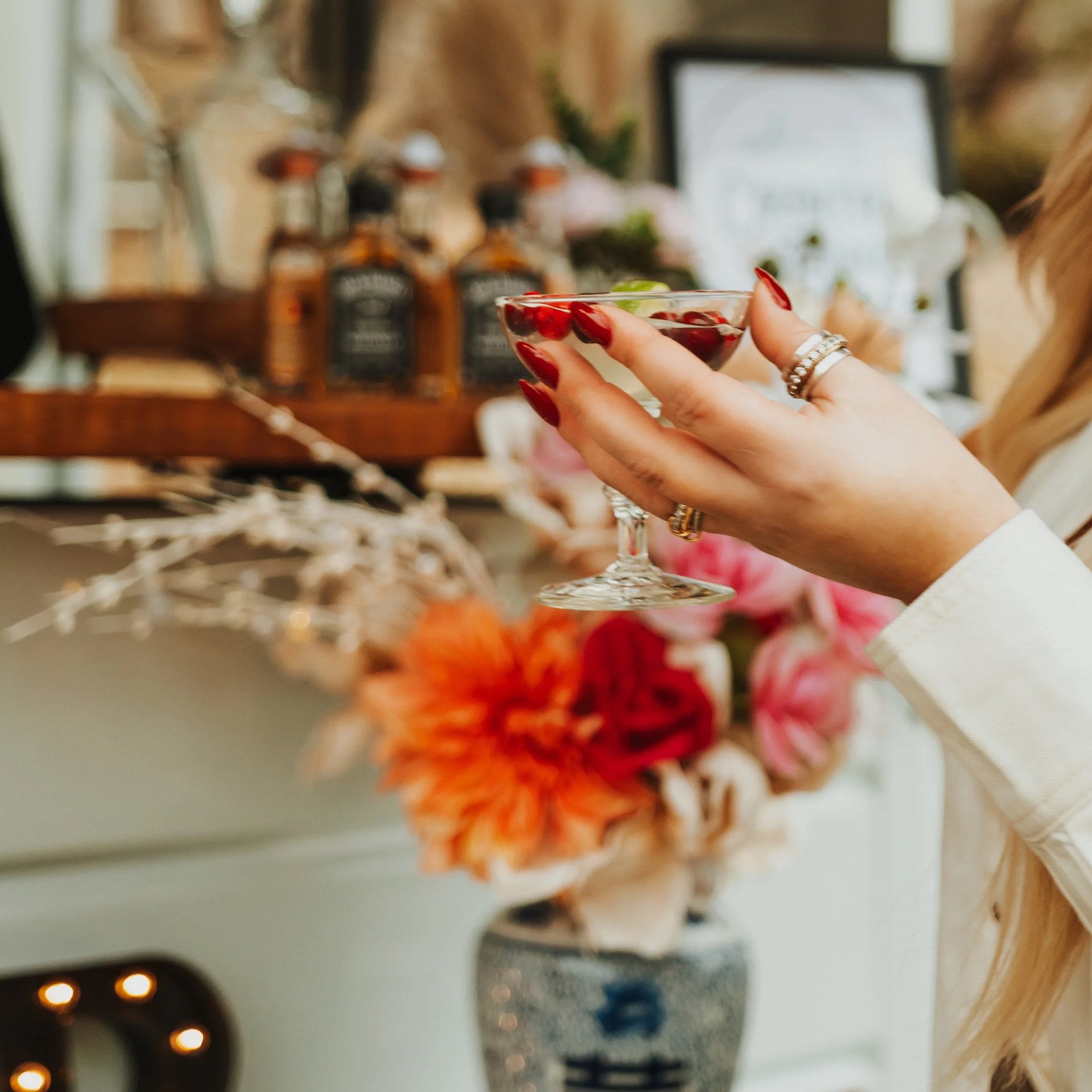 Person holding a cocktail glass with cranberries and lime, decorated with red nail polish and rings, at a festive gathering with flowers and Jack Daniel's bottles in the background.