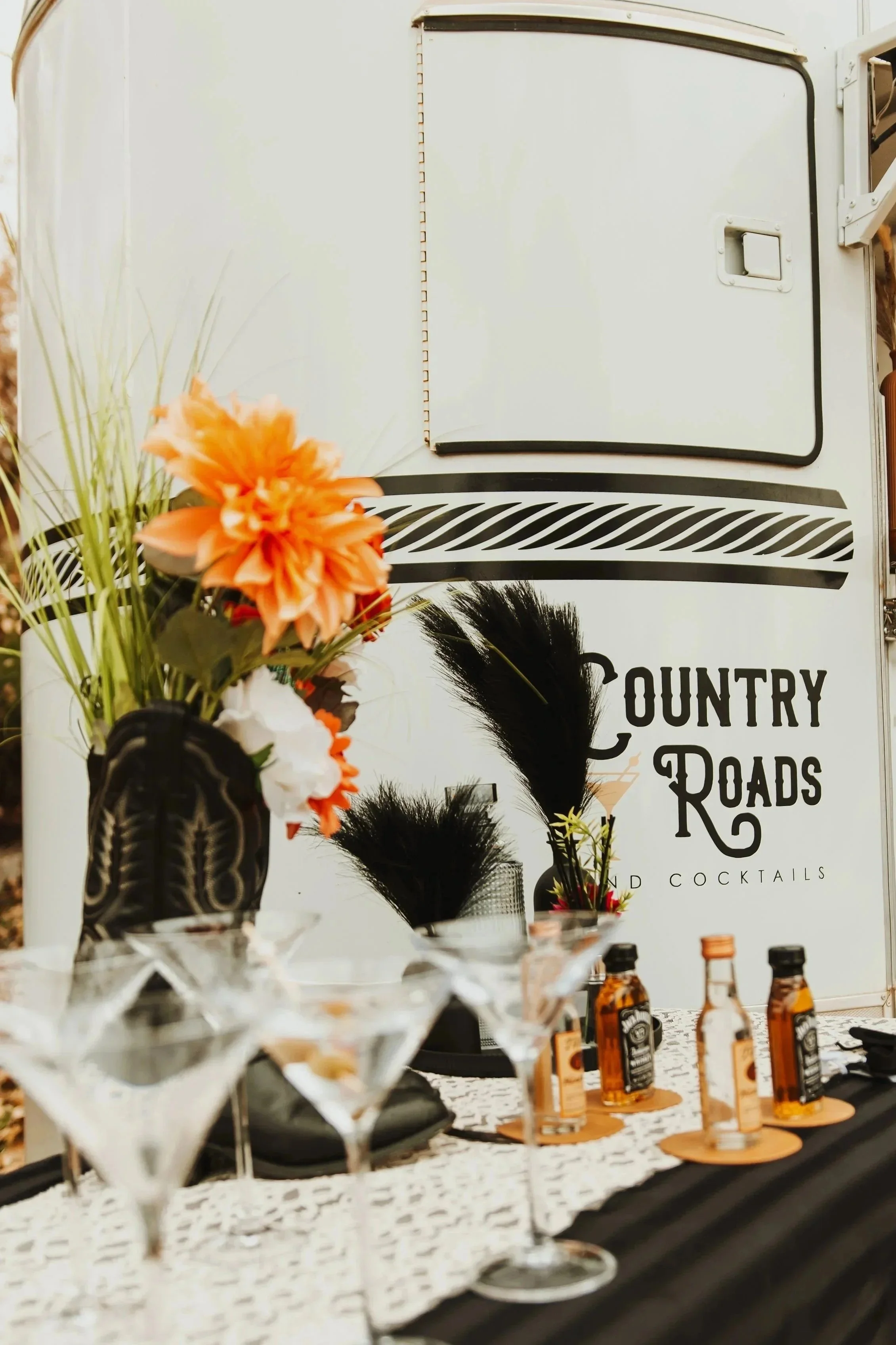 A table set for drinks in front of a food truck labeled 'Country Roads'. The table has cocktail glasses, bottles of Jack Daniel's whiskey, and decorative feathers and flowers.