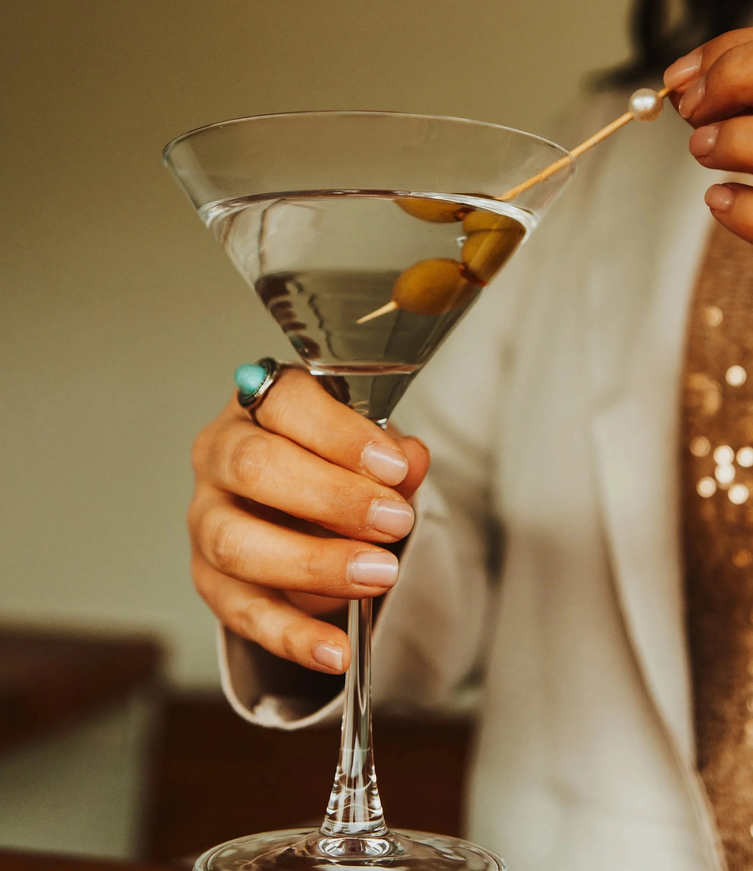 Close-up of a hand holding a martini glass with olives and a person garnishing the drink with an olive on a toothpick.