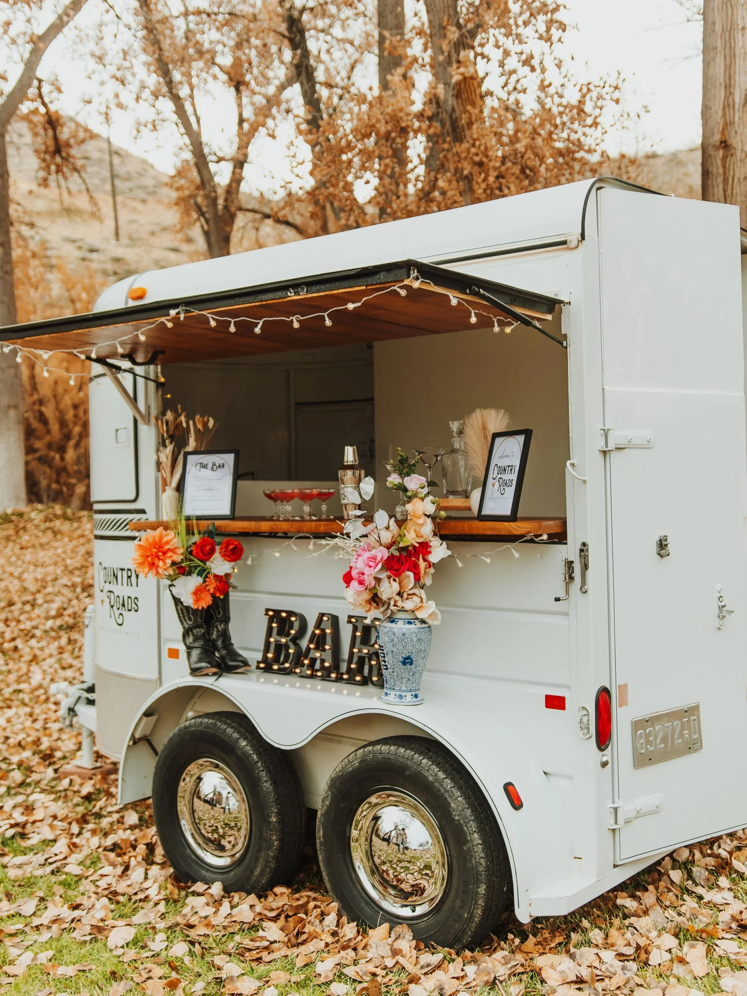A white mobile bar truck decorated with flowers, fairy lights, and a black Mrs. sign in an outdoor setting with autumn leaves and trees.