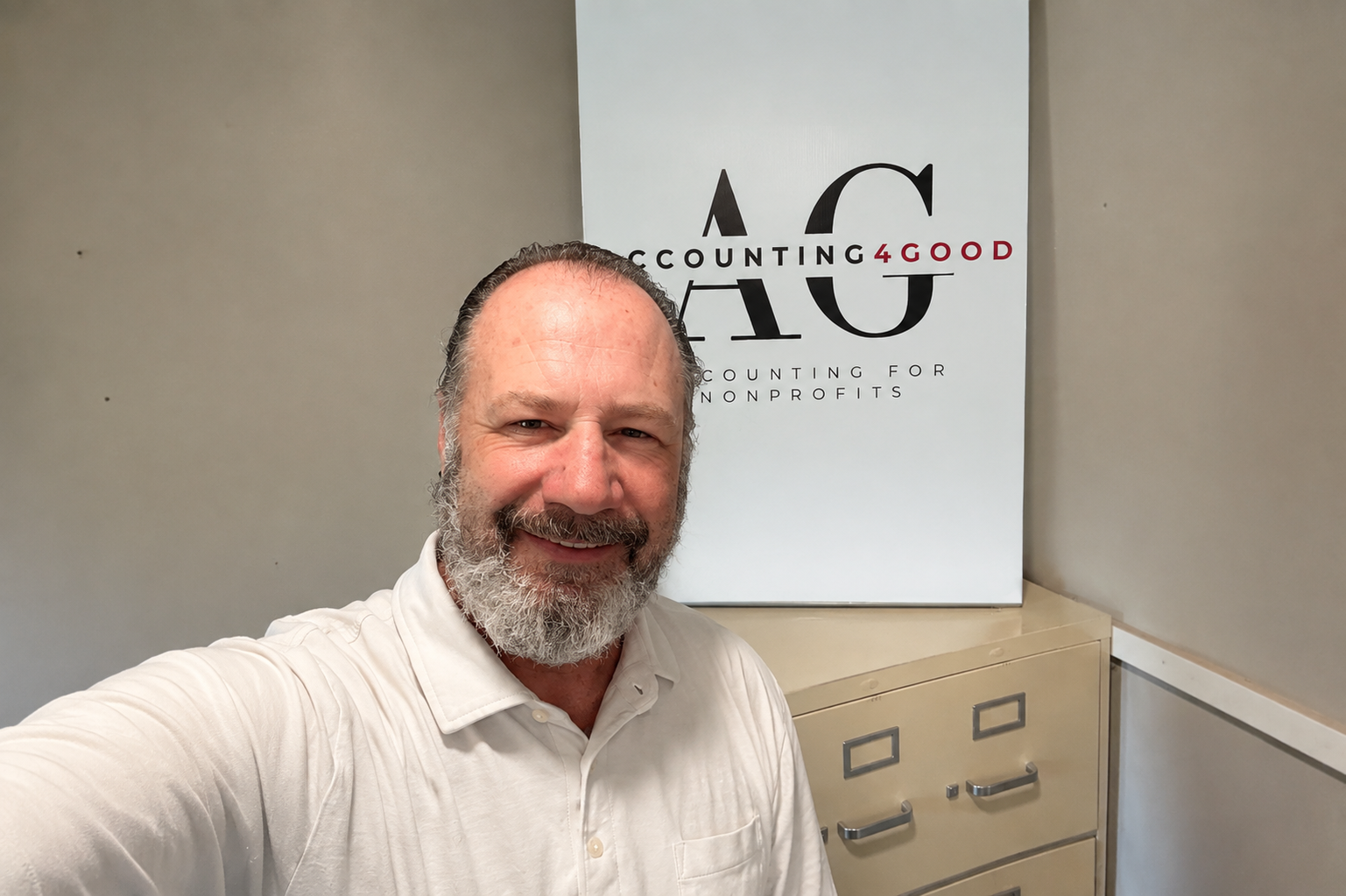 A smiling middle-aged man with a beard taking a selfie in front of a sign that reads 'Accountings4Good, Accounting for Nonprofits'. He is wearing a white collared shirt and standing in an office with beige cabinets.