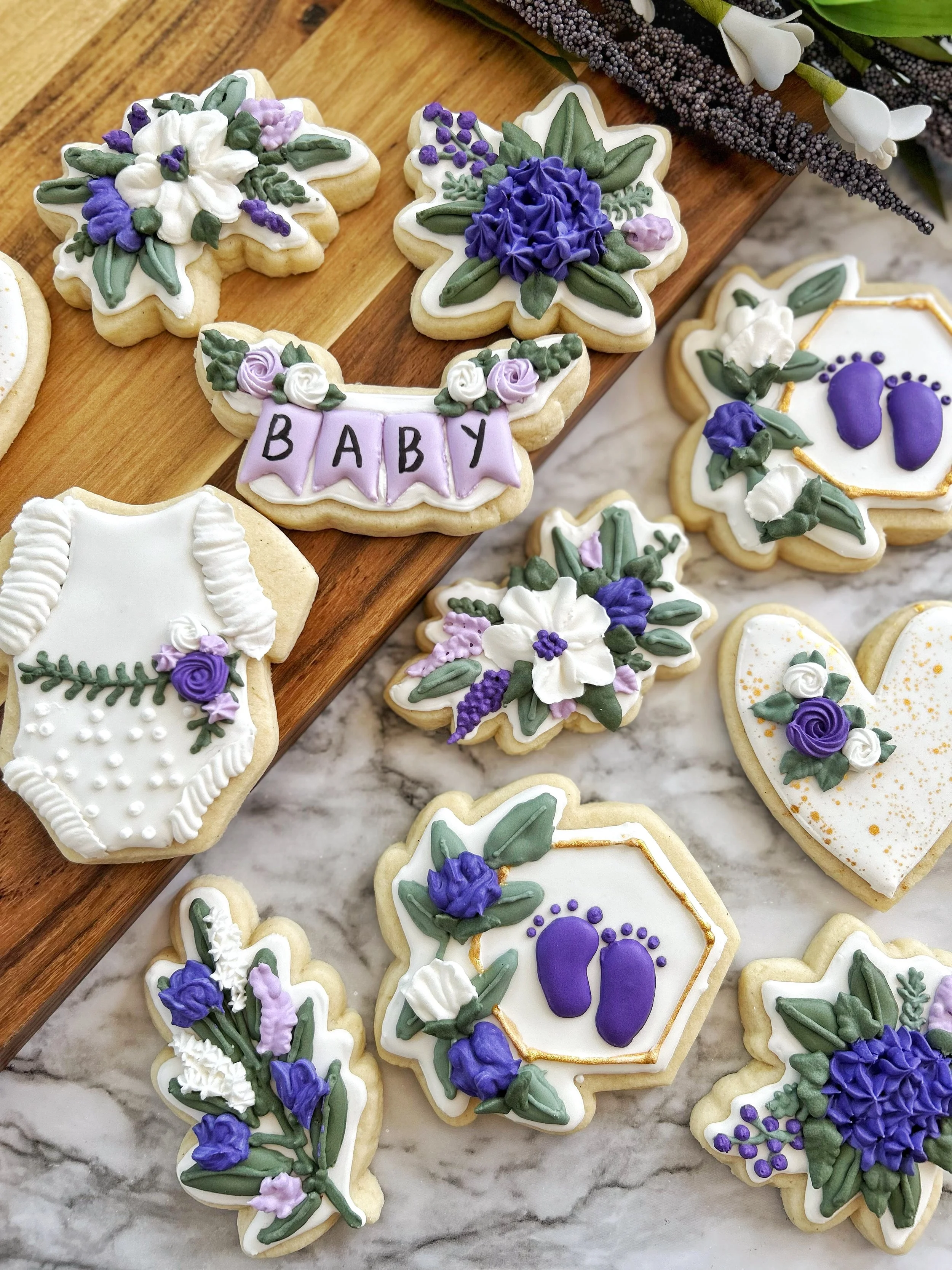 Decorated cookies with floral and baby themes, including one cookie with the word 'BABY' on purple banners and lavender, purple, and white icing flowers, set on a marble surface with a wooden board and flower arrangement.