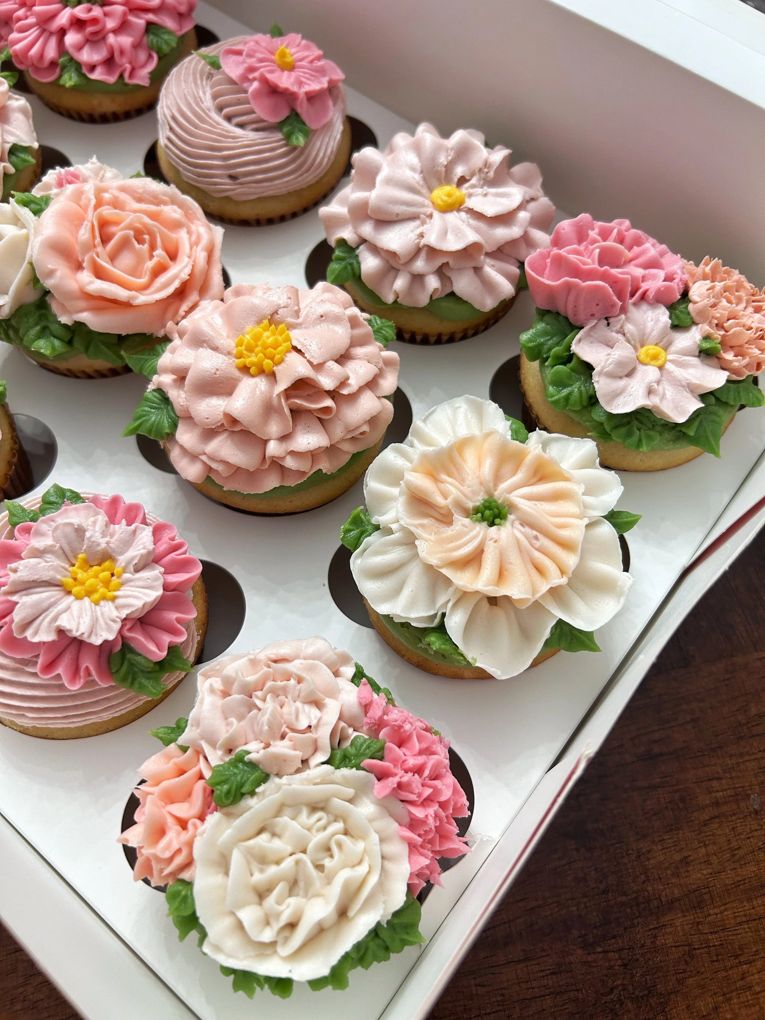 A tray of decorated cupcakes with floral icing designs in pink, white, and cream colors with green icing leaves.