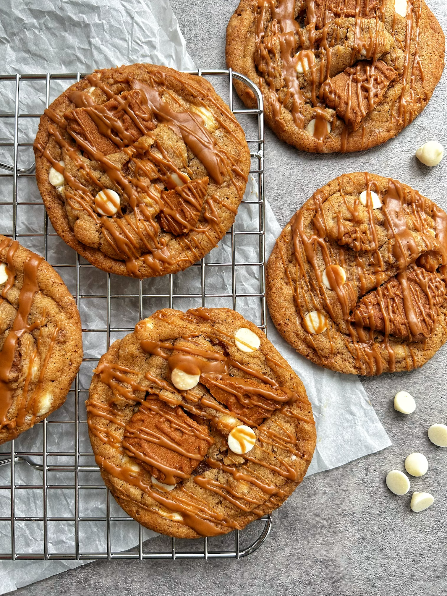 Several large cookies with white chocolate chips drizzled with caramel, placed on a cooling rack and a gray surface. White chocolate chips are also scattered nearby.