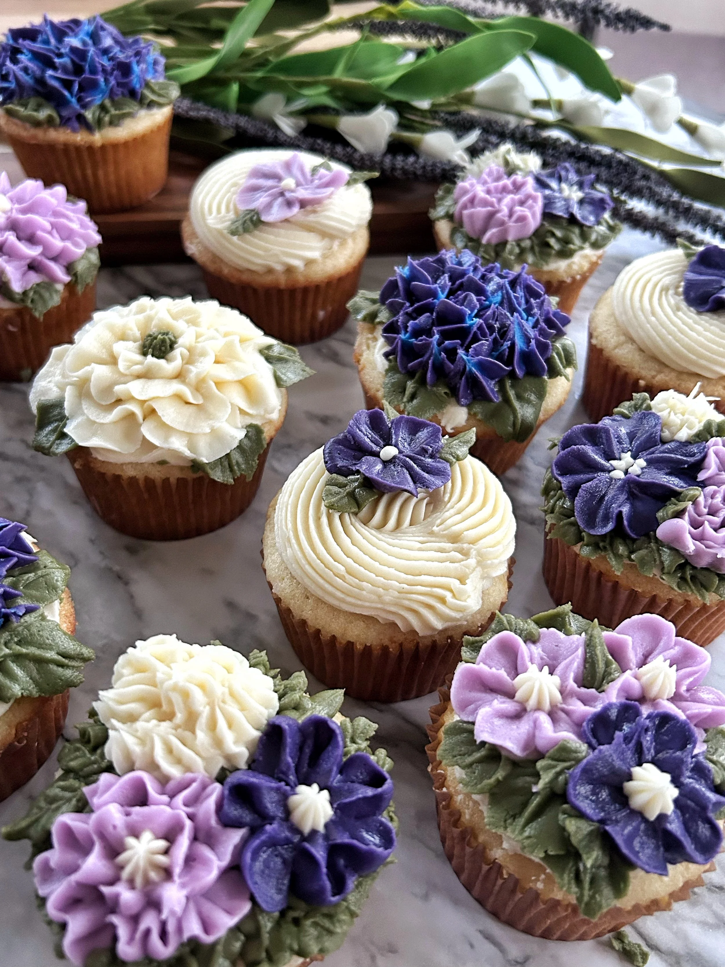 Assorted cupcakes decorated with floral icing in purple, white, and pink colors, topped with green leaves, arranged on a marble surface with a bouquet of white and purple flowers in the background.