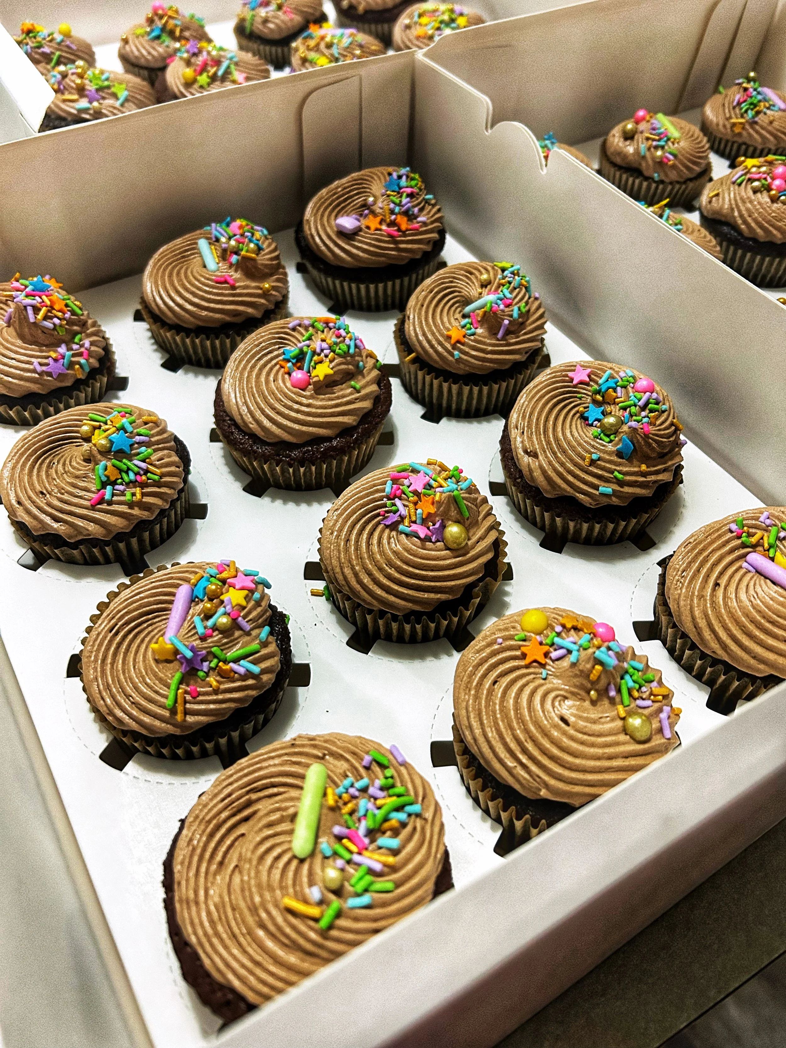 A white box holding multiple chocolate cupcakes with brown frosting, decorated with colorful sprinkles, star-shaped, round, and elongated candies.