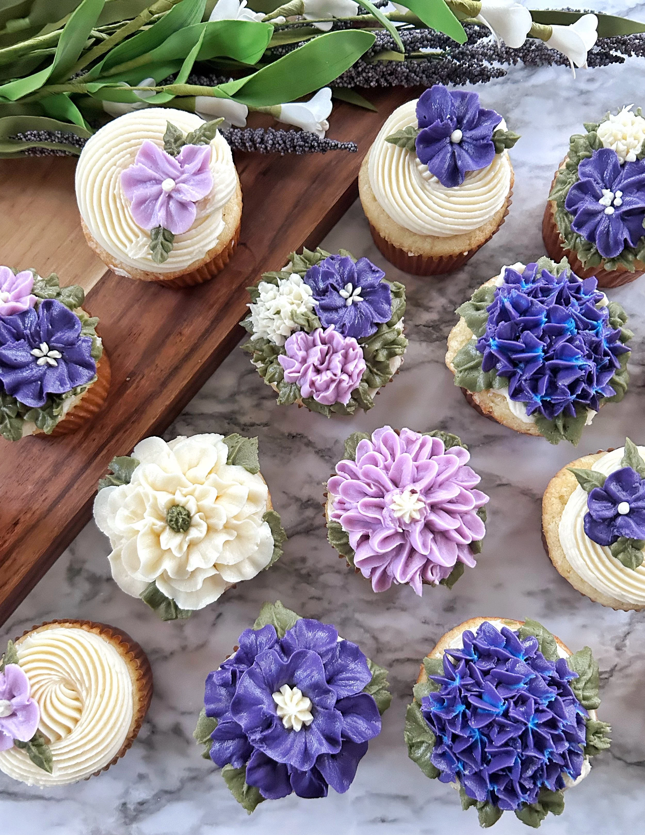 Assorted cupcakes decorated with purple, white, and lavender flowers made from icing, with some green leaves, arranged on a marble surface with a wooden board and floral arrangements nearby.