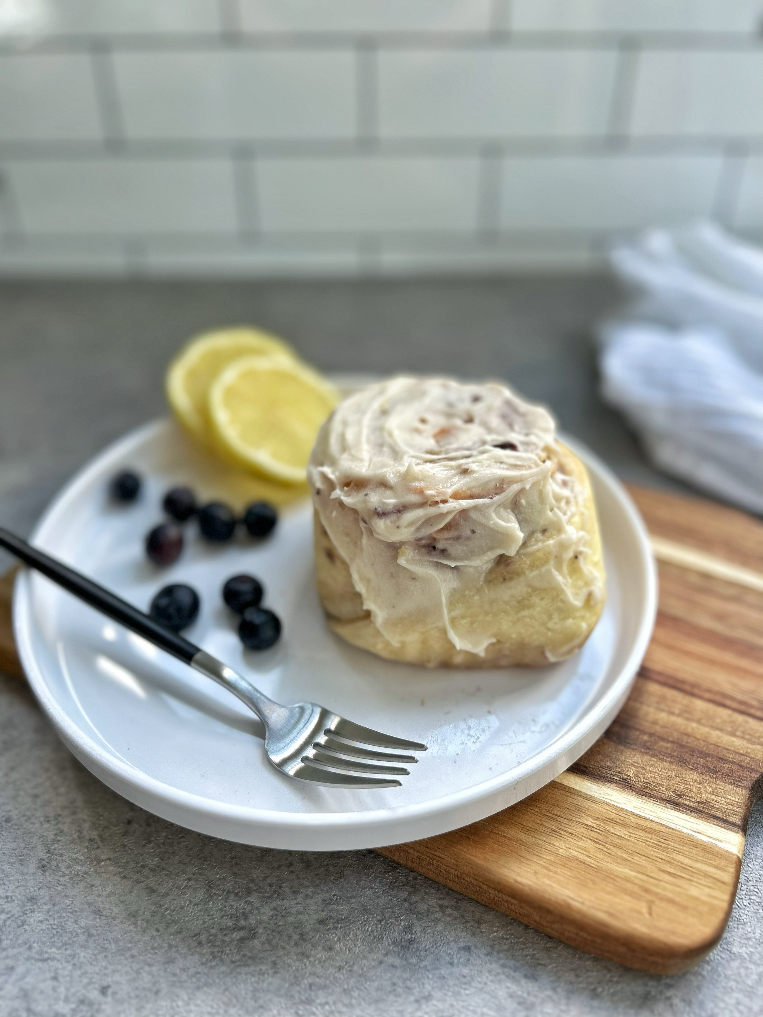 A plate with a piece of lemon-flavored sweet roll with cream cheese frosting, garnished with lemon slices and blueberries, on a white plate on a gray surface next to a wooden cutting board.