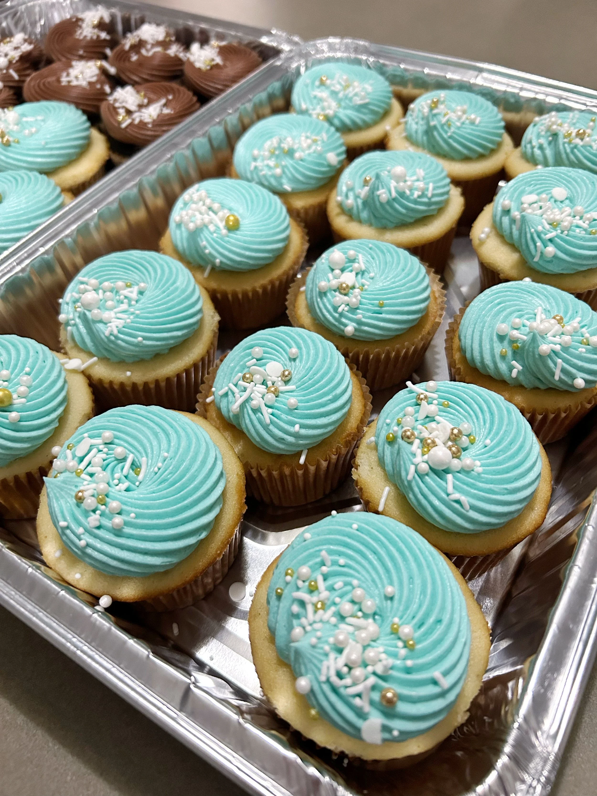 A tray of cupcakes with blue frosting and white, gold, and pearl decorations, alongside a tray of chocolate cupcakes with chocolate frosting and coconut flakes.