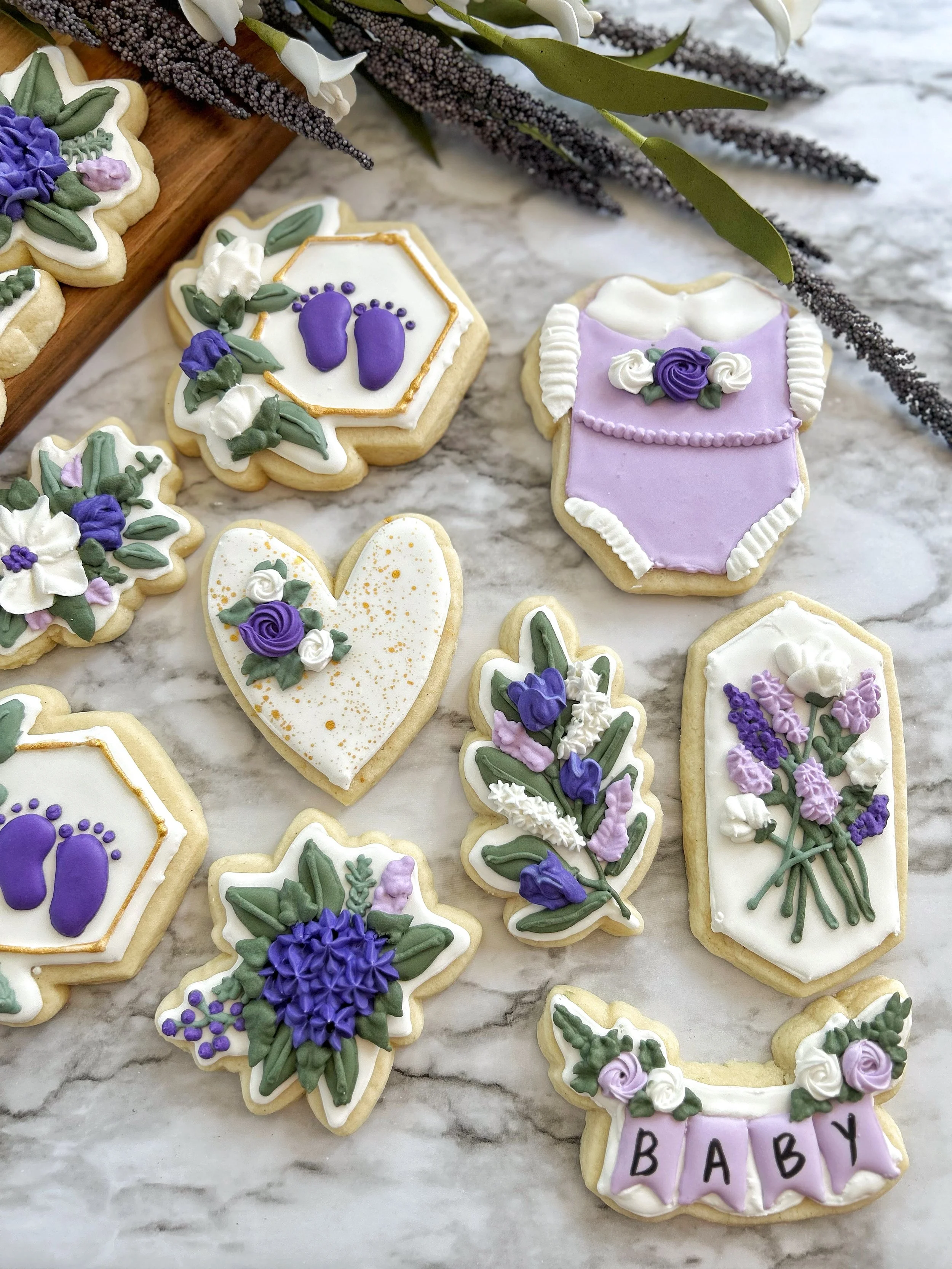 Decorated cookies with lavender and purple flowers, including baby-themed designs and a banner with 'BABY' written on it, arranged on a marble surface with floral accents in the background.